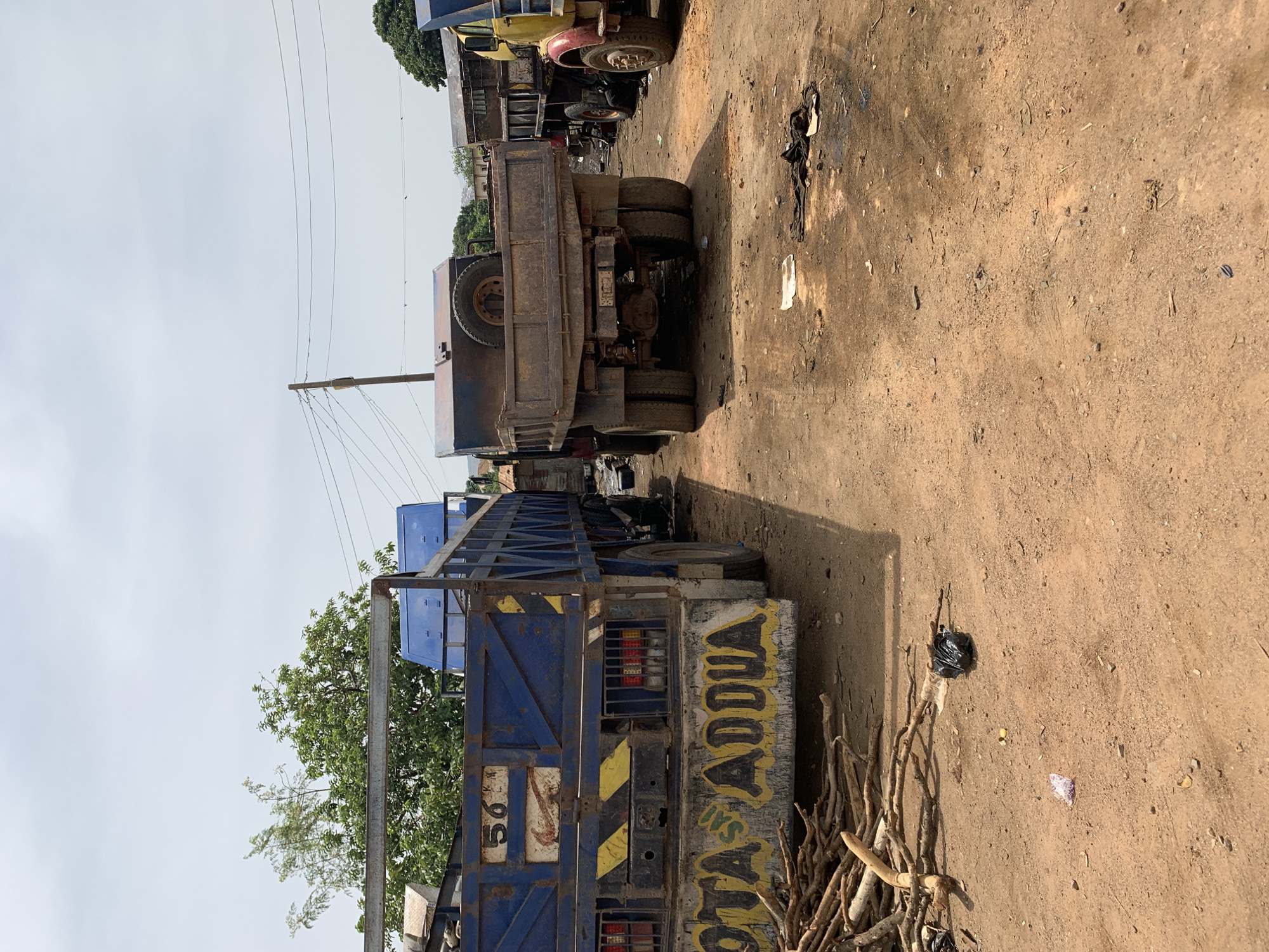 The image depicts a street in Rafin Par, Nigeria, with several trucks parked along a dirt road. A blue truck with "OTA ADDDA" painted on the back is visible in the foreground, with a pile of branches nearby. Behind it is another truck, its rear facing the camera. Further down the road, other trucks are parked, including a yellow one. Overhead, utility poles and wires are visible against a cloudy sky. The ground is dry and dusty, with scattered debris. No people are visible in the frame.