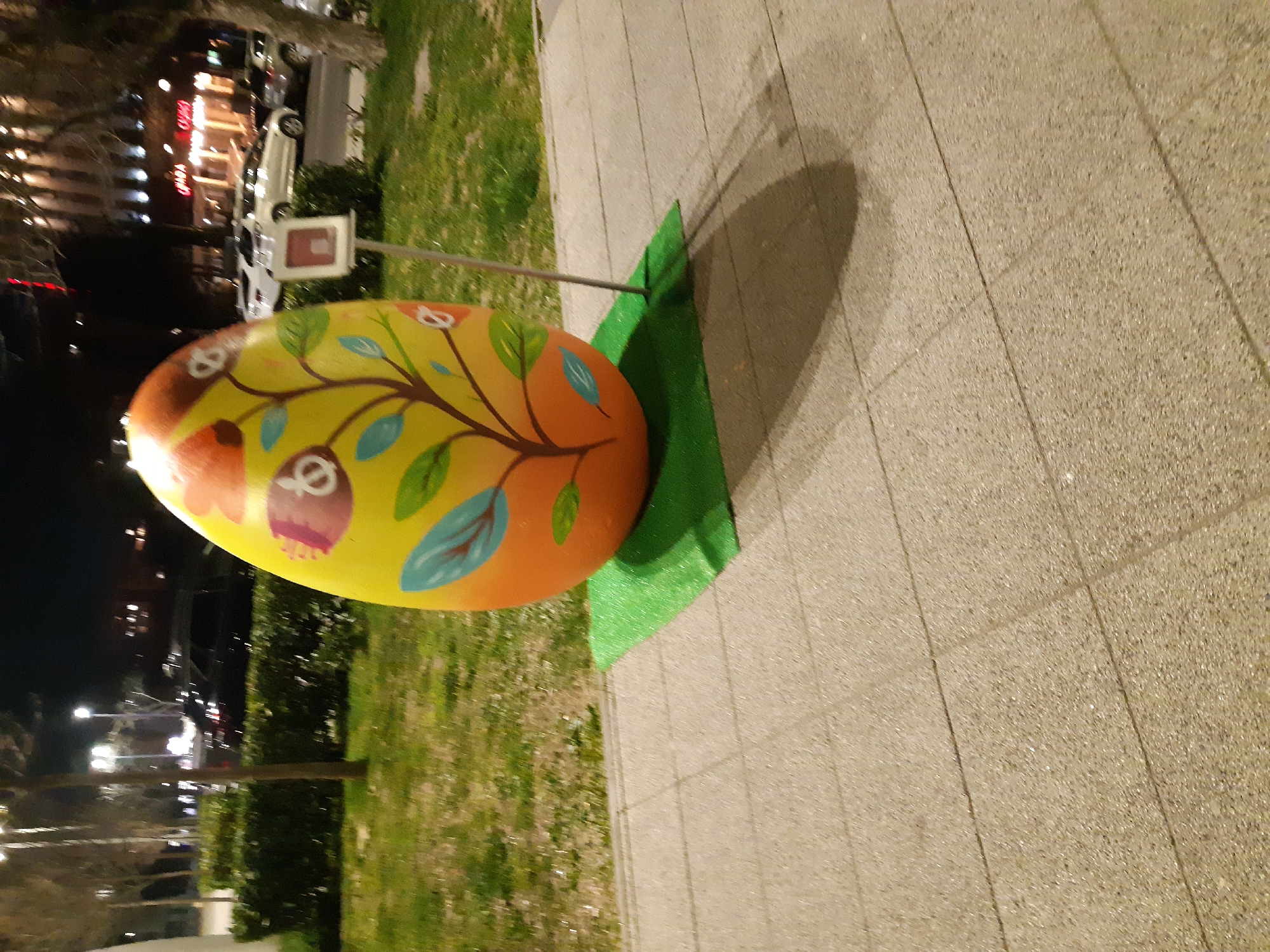 A brightly painted, oversized Easter egg stands on a patch of green artificial turf adjacent to a paved walkway. The egg is yellow and orange with a stylized tree branch painted on it, bearing leaves and decorative egg-shaped ornaments. A metal support rod connects to the egg. The backdrop includes green grass, bushes, and trees. In the distance, buildings with illuminated signs and lights suggest an urban setting, likely Plovdiv, Bulgaria. The scene is likely photographed at night or dusk, with artificial lighting illuminating the egg and its surroundings.