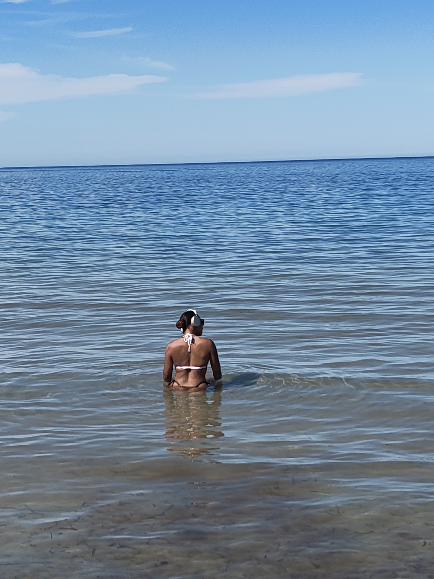 The image depicts a serene coastal scene featuring an individual immersed in the shallow, clear waters of the sea. The person, viewed from behind, appears to be female and is wearing a white bikini. White over-ear headphones are visible on her head. She is facing away from the camera, looking out towards the vast expanse of the ocean and the distant horizon.
The setting is bright and sunny, indicative of daytime. The sky is a clear blue with a few subtle white clouds, particularly visible in the upper sections of the image. The sea surface is relatively calm, displaying gentle ripples. In the foreground, the water is shallow enough to reveal the sandy or rocky seabed, which has some visible seaweed or marine vegetation. Further out, the water takes on a deeper blue hue as it extends towards the horizon.
The overall atmosphere suggests peace and relaxation, with the individual seemingly enjoying the tranquility of the water and the view. No text is visible in the image. The location is Dénia, Spain.