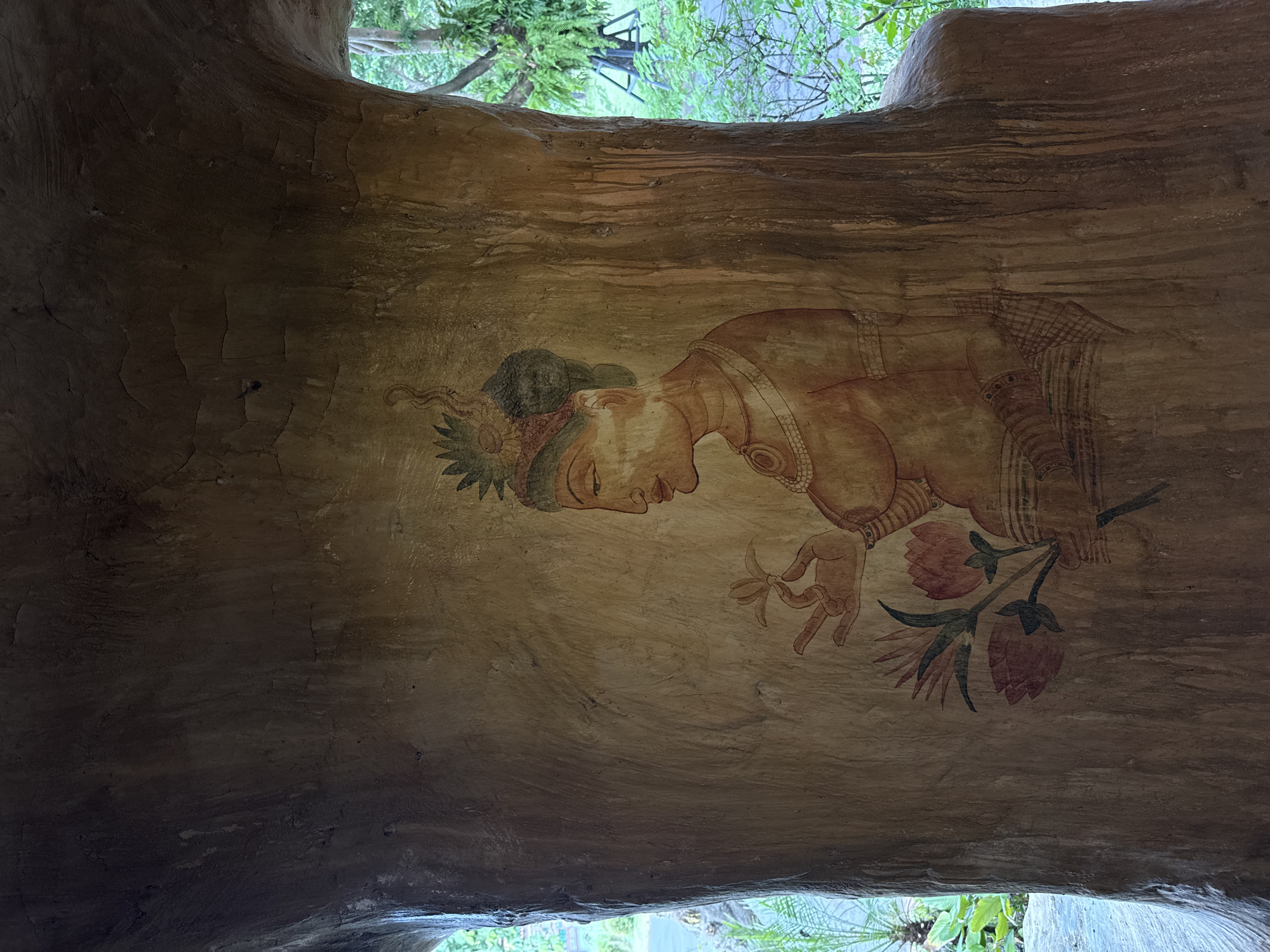 The image shows a mural painted on a textured, light brown, tree-like structure, possibly part of an architectural feature or natural formation. The mural depicts a profile of a figure, likely female, with dark hair styled in an updo adorned with green foliage and a red band. The figure has prominent facial features, including a straight nose, almond-shaped eye, and full lips. They are wearing jewelry, including a necklace with circular pendants and armbands. The figure's right hand is raised, holding a delicate, light-colored flower. Below the figure, a cluster of three pink lotus flowers with green stems and leaves is painted. The artwork is rendered in earthy tones of brown, red, and green. The structure itself has a rough, uneven surface with visible cracks and streaks, suggesting age or an organic texture. The background visible through an opening in the structure is a lush green outdoor environment with trees and foliage. The location is identified as Bang Pu Subdistrict Municipality, Thailand.