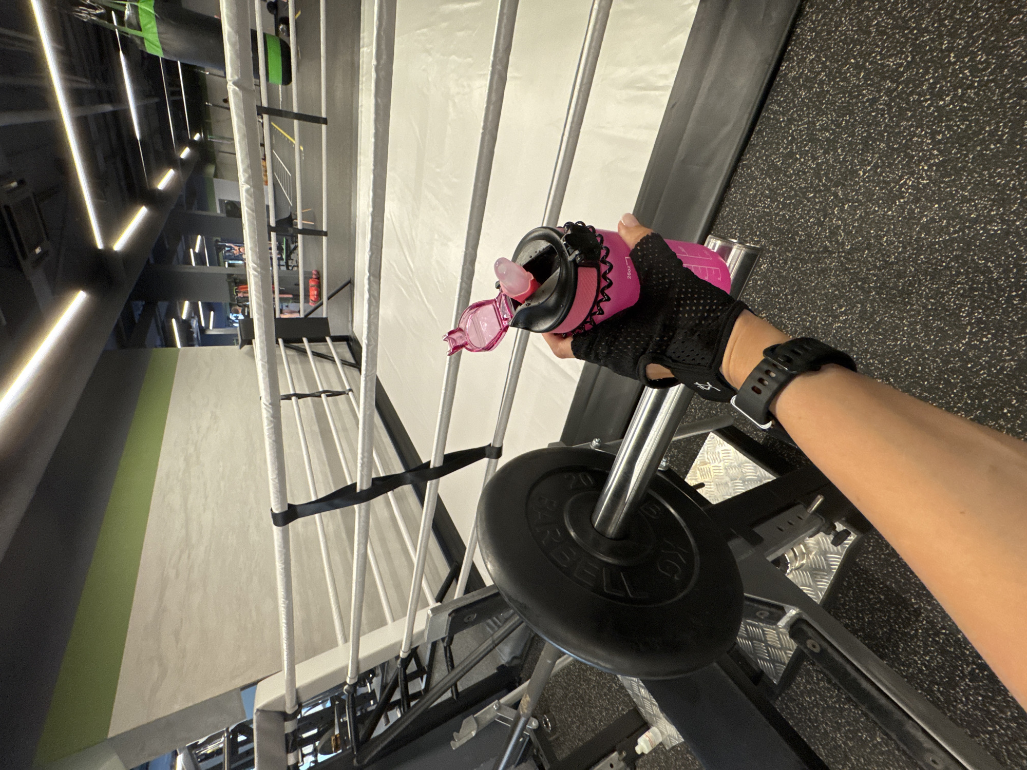 The image depicts a close-up of a person's hand, wearing a black fingerless glove with a textured grip, holding an open pink water bottle. The hand also wears a black smartwatch. The water bottle is positioned above a weight plate, which is part of a barbell situated on a weight machine. The weight plate has visible text that appears to read "20 KG" and "BARBELL".
The background shows a gym setting. To the left, there are rows of vertical metal bars, possibly part of a rack or storage unit, with white padding or covering. Further in the background, there are gym lights and equipment, with some signage visible. The floor is a speckled black rubber mat. The overall impression is a moment during a gym workout. The lighting suggests it is indoors, and there are no indicators of time of day or weather. The context provided indicates the location is Sochi, Russia, but this is not directly discernible from the image itself. No specific emotions are conveyed by the image.
