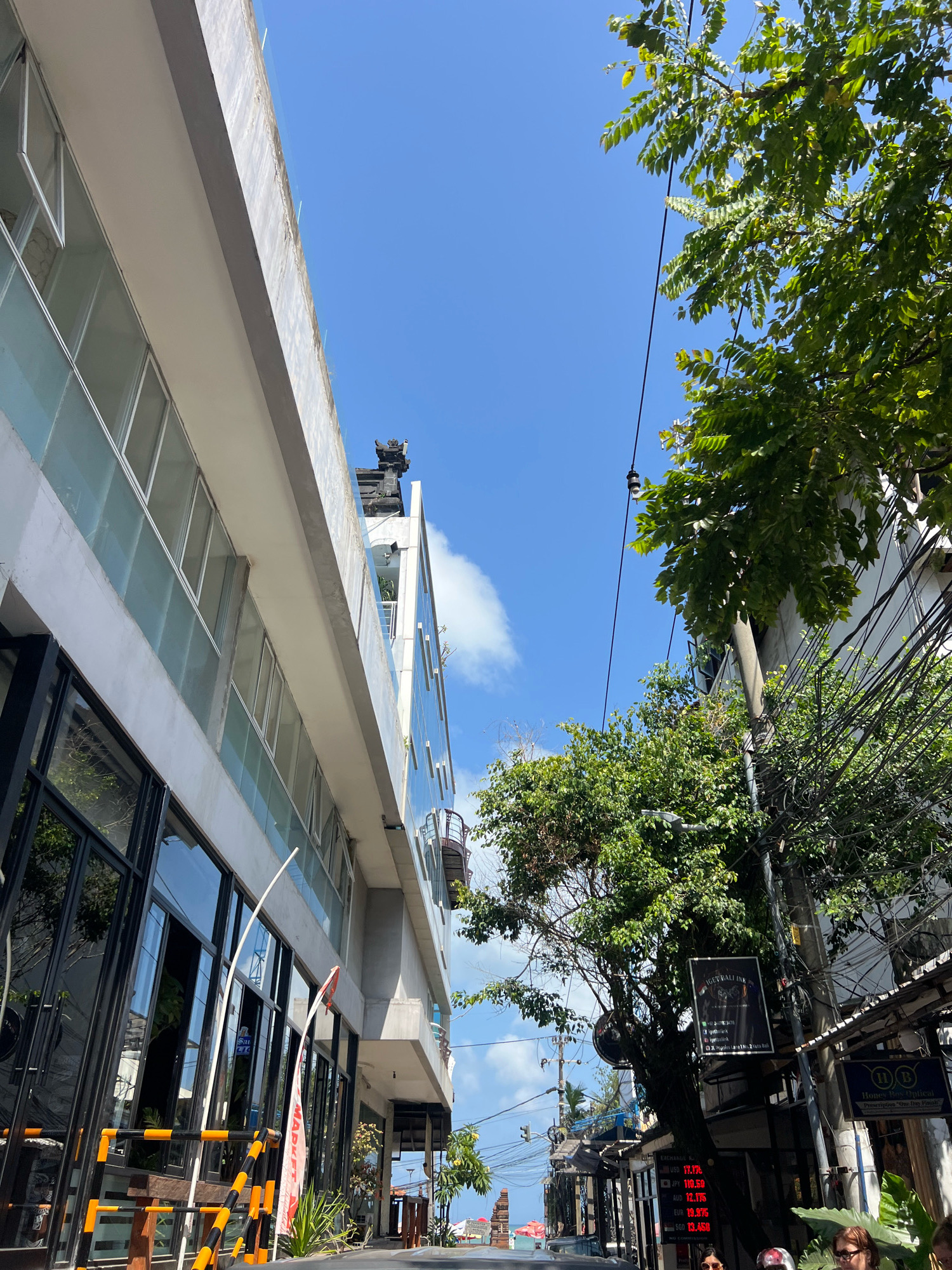 In Kuta, Indonesia, a street scene unfolds under a bright, clear blue sky with sparse white clouds. The perspective is from ground level, looking upwards and diagonally. To the left, a modern multi-story building features a sleek facade of white and grey concrete, with large, floor-to-ceiling windows with black frames reflecting the surroundings. The building's upper edge is visible, with a protective railing and some minor wear. On the right side of the frame, a different building, possibly older, displays a more cluttered facade with numerous electrical wires strung across its exterior. A large, leafy green tree dominates the right side, its branches reaching into the sky and partially obscuring the building. Several signs are attached to poles and buildings. One sign, a black rectangle with white text and graphics, appears to be an advertisement. Further down the street, other signs and storefronts are visible, including one for "Honest Eye Optical." The street level is not clearly visible, but there are hints of traffic barriers and some vegetation. In the distance, a prominent Balinese-style architectural element, possibly a temple gate or tower, can be seen. The overall impression is one of a bustling commercial street in a tropical location.