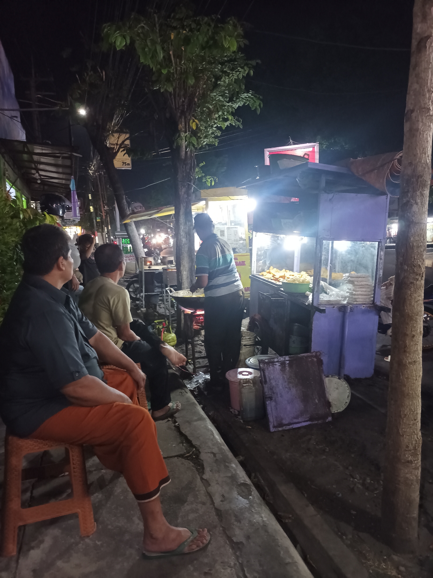 A nighttime outdoor scene in Nganjuk, Indonesia, depicts a street food vendor and customers. A vendor, wearing a striped shirt and dark pants, is actively cooking at a food stall that features glass display cases filled with fried food items. Several individuals are seated on stools, observing the vendor or waiting for their orders. One person in the foreground is wearing a dark shirt and orange pants, while another is in a tan shirt and dark pants. The environment includes trees, streetlights, and other food stalls visible in the background, suggesting a busy street market or food alley. The ground appears to be paved, with some debris and containers present.