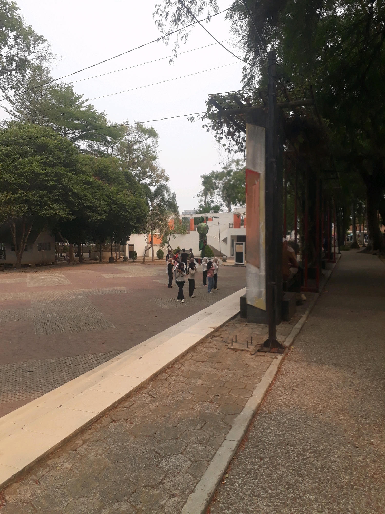 The image depicts an outdoor scene, likely a public square or park area in the city of Pangkalpinang, Indonesia. In the foreground, a paved walkway with a raised curb runs along the right side of the image. The walkway is composed of textured grey material with visible aggregate. Beyond the curb, a wider paved area, possibly a plaza, features a repeating pattern of red and grey interlocking bricks.
On the left side of the plaza, a low, light-colored building with a dark roof and visible windows is partially obscured by trees. Further back, a white building with orange architectural accents is visible. A decorative, green, bulbous sculpture stands near this building.
A group of approximately eight to ten individuals, appearing to be young people, are gathered in the middle ground of the plaza. They are casually dressed, some wearing headscarves, and appear to be interacting with each other or observing something.
The background is dominated by a dense canopy of trees, with varying shades of green foliage. Power lines are visible crisscrossing the sky, which is overcast with a light grey hue. On the far right, a line of dark poles with a lattice structure overhead suggests a covered walkway or trellis, with some vegetation growing on it. The overall impression is of a slightly hazy, diffused daylight.