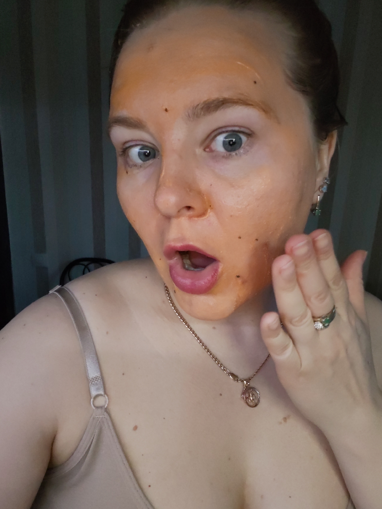 A woman with a facial mask is shown in an indoor setting. The woman has light skin, blue eyes, and reddish-blonde hair pulled back. An orange facial mask is applied to her face, with a few dark spots visible within the mask. She has a surprised expression, with her mouth open and eyes wide. Her right hand is raised near her face, with a ring on her finger. She is wearing a beige tank top, and a gold necklace with a pendant is visible around her neck. Earrings are present in her ear. The background consists of vertical striped curtains.
The image was not taken in the city of Izhevsk, Russia.
