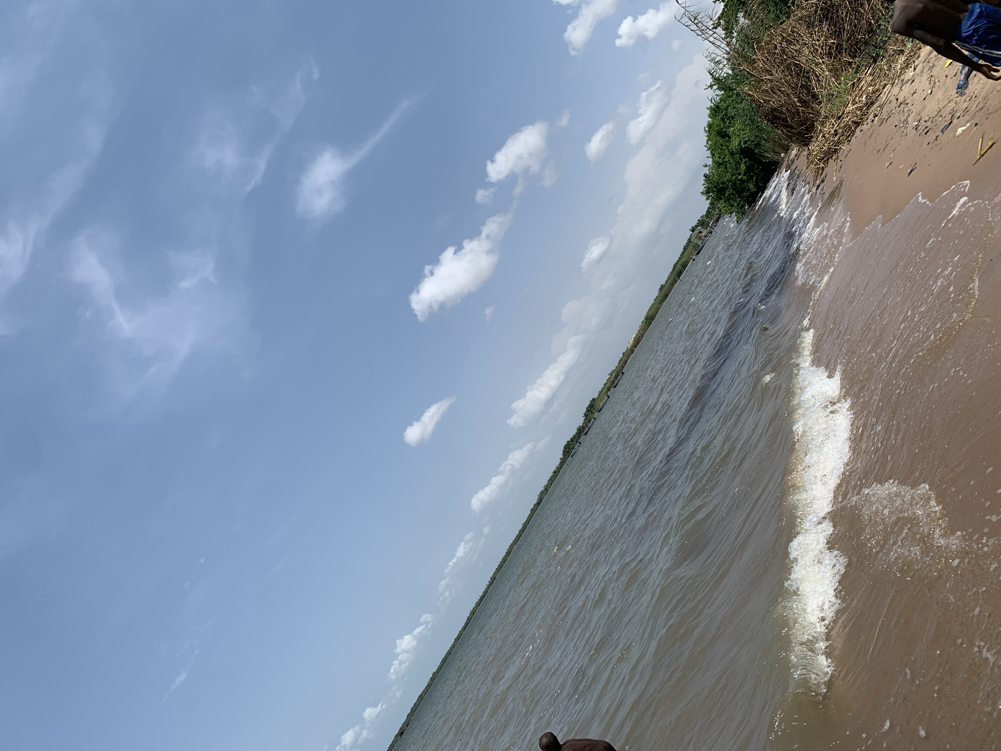 The image depicts a beach scene, likely in Lekki, Nigeria, given the prompt's specification. The shoreline curves from the upper right to the lower center of the frame. Waves are breaking on the sandy beach. Dense vegetation borders the beach on the upper right. The water is a murky color. The sky is blue with scattered white clouds. Parts of two people are visible on the upper and lower right, the upper person wearing blue shorts.