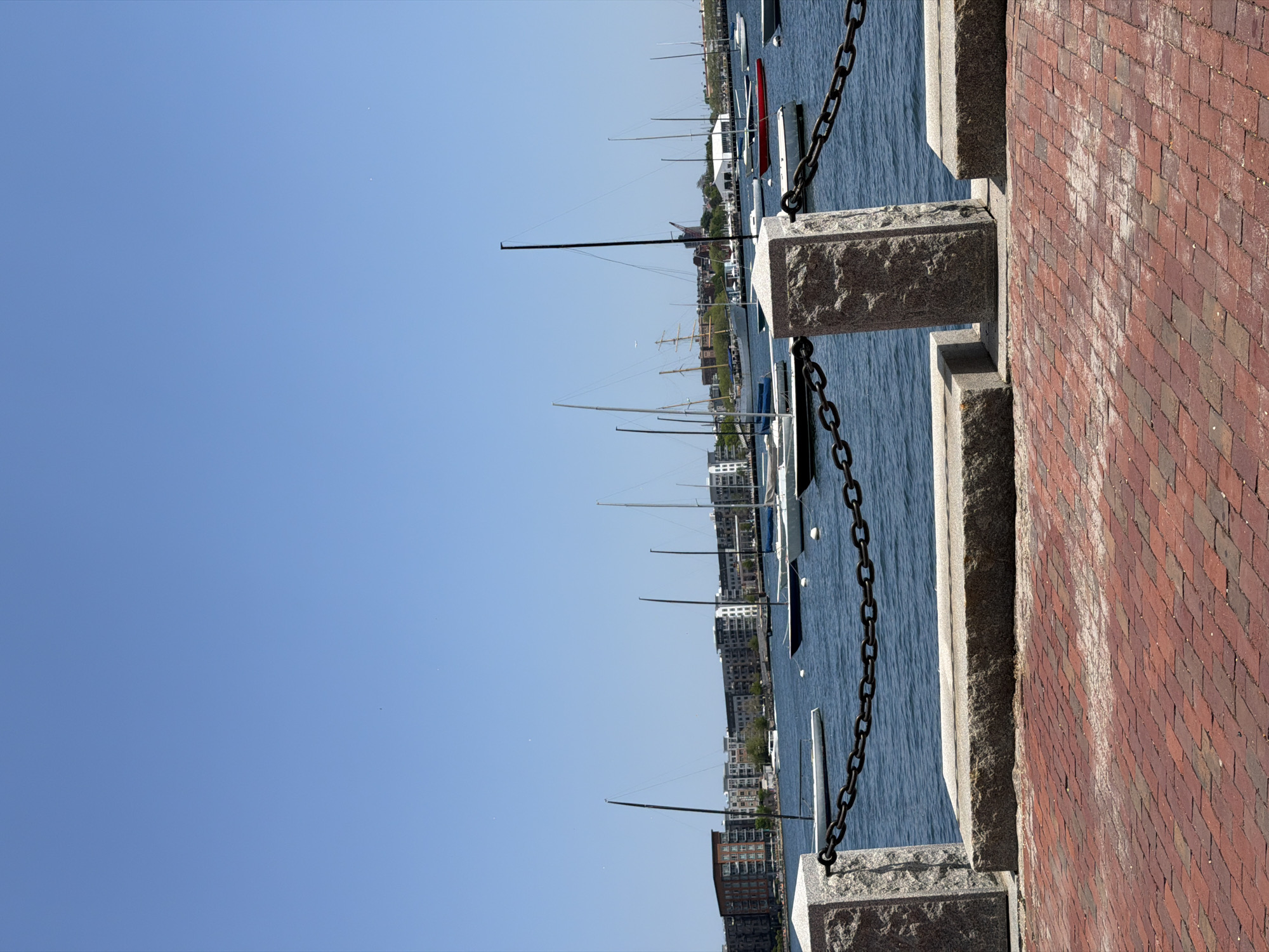 This is an outdoor shot, taken in the city of Boston, Massachusetts, USA. The image shows a waterfront scene with boats and buildings in the background. A stone and brick wall runs vertically on the right side of the image, with a black metal chain suspended between the stone pillars. The water is visible in the middle, with various boats and masts reaching towards the clear blue sky. In the distance, buildings line the shore. The overall composition gives a sense of being on a waterfront promenade, looking out at the harbor.
