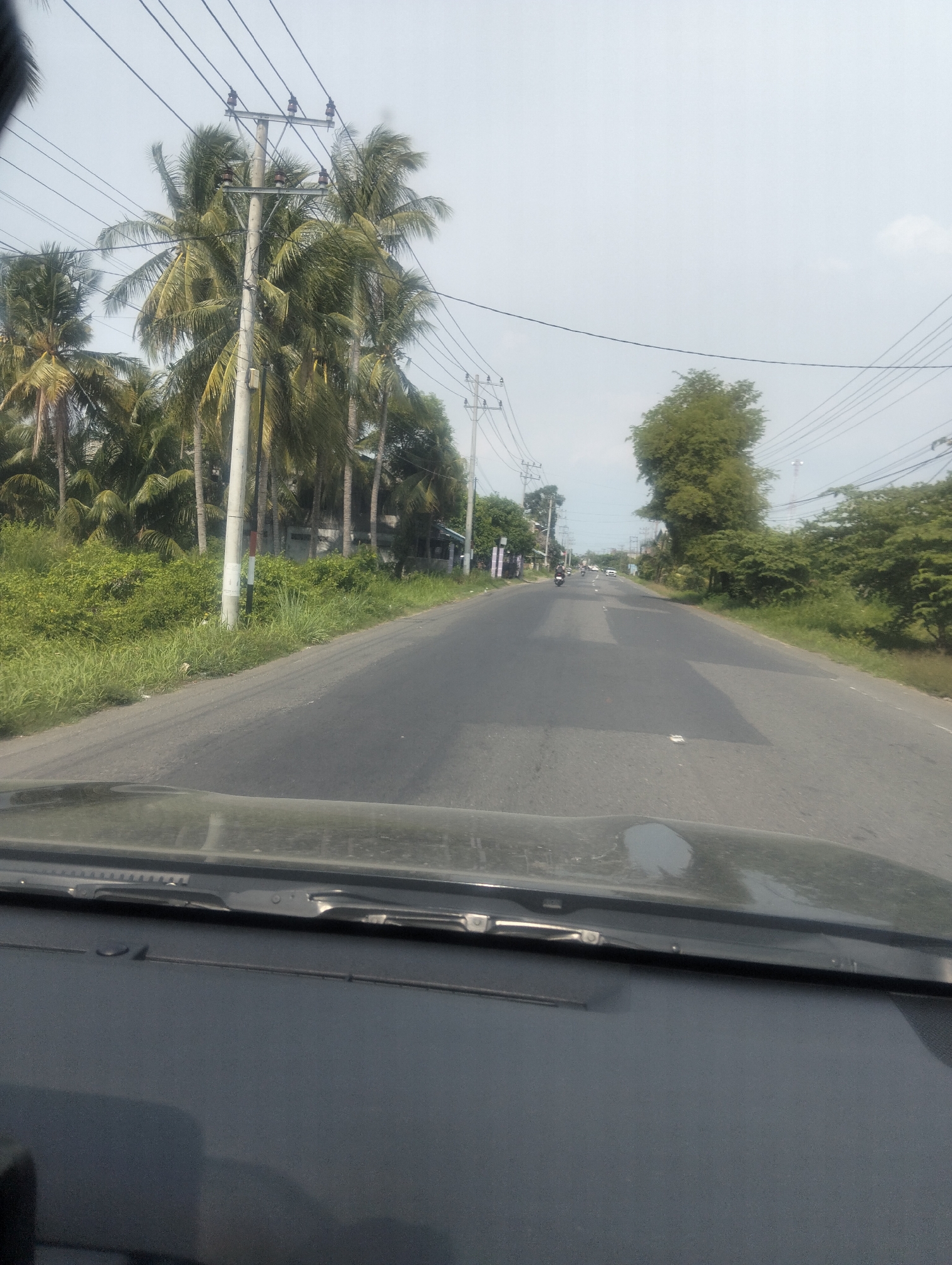 The image shows a view from inside a vehicle, looking out onto a paved road in Cotbadatunong, Indonesia. The road is flanked by lush green vegetation, including numerous palm trees. Overhead, power lines are strung between utility poles. Several motorcycles and a few cars are visible on the road, indicating ongoing traffic. The sky is overcast with a hazy, light gray appearance. The foreground of the image is dominated by the dark interior of the vehicle, including the dashboard and windshield.