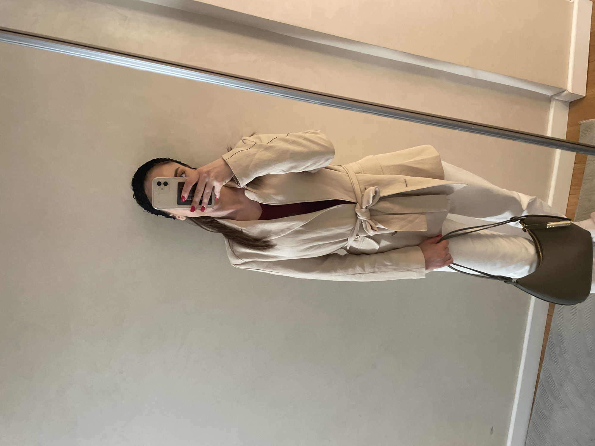 A woman stands indoors, angled slightly to the left, taking a mirror selfie. She wears a black headband, a tan belted jacket, and white pants. Her nails are painted red, and she holds a light-colored smartphone in her left hand. She also has a dark green purse slung over her shoulder. The background is a light beige wall with a mirror framed in metal. There is a glimpse of a wood floor and rug on the right side of the image. The setting is most likely an interior space within a home or apartment. The location is not evident from the image, and there is no way to know if this location is within Varna, Bulgaria.