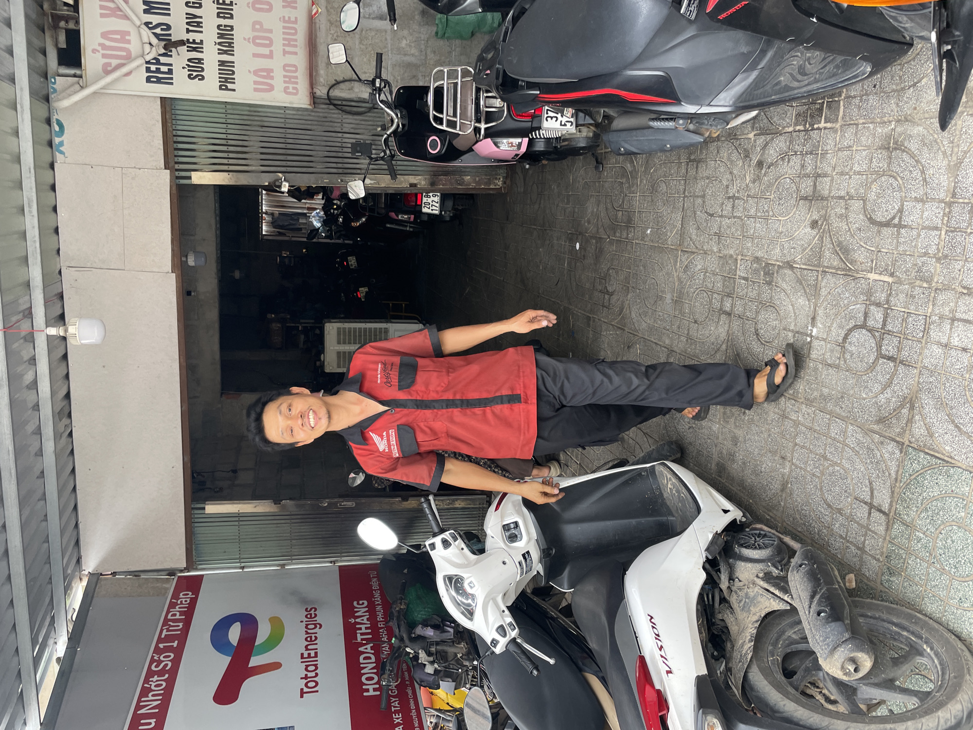 The image depicts a mechanic in Phu Thuy Ward, Vietnam, standing outside a motorcycle repair shop. He is wearing a red Honda uniform and smiling. The shop's sign advertises repairs, tire patching, and motorcycle rentals. Several motorcycles, including a white Honda Vision and a black scooter with pink details, are parked nearby. A TotalEnergies/Honda Thang sign is also visible. The shop itself is open, with glimpses of more motorcycles inside. The ground is paved with patterned tiles.