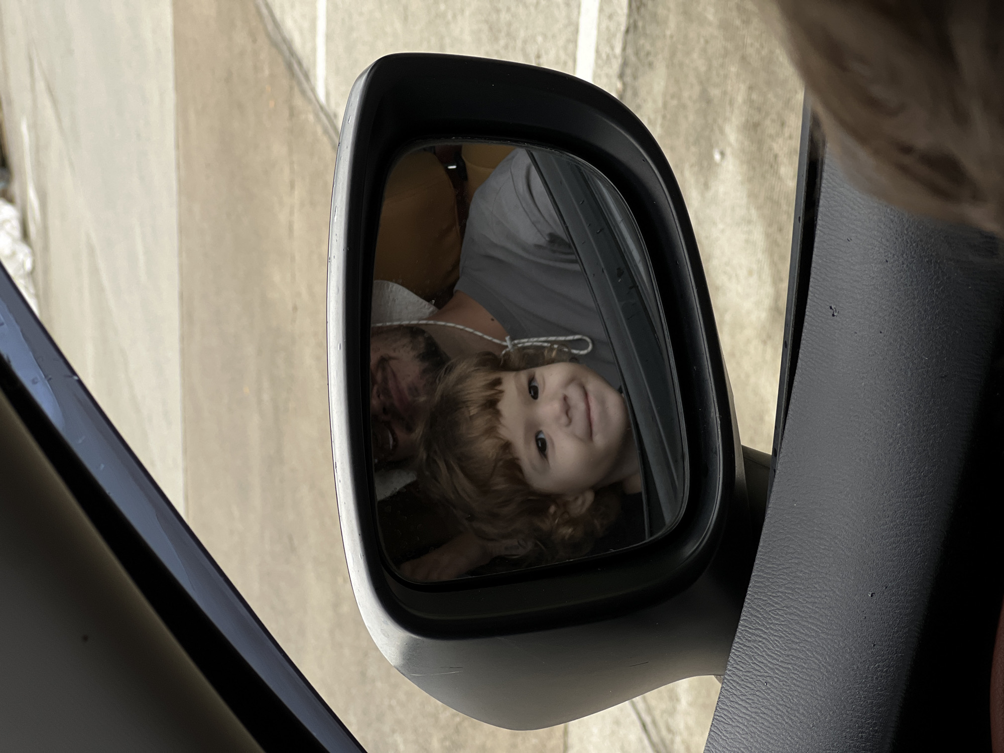 The image shows the reflection of a child and an adult in the side mirror of a vehicle. The child, with curly brown hair, is positioned in the lower right quadrant of the mirror's reflection and is smiling. An adult, possibly wearing a grey shirt, is visible above and to the left of the child, with their face partially obscured. A white mask appears to be hanging around the adult's neck. The car's black interior panel is visible in the foreground. Outside the mirror, there is a glimpse of a grey urban landscape, suggesting a location in a city.