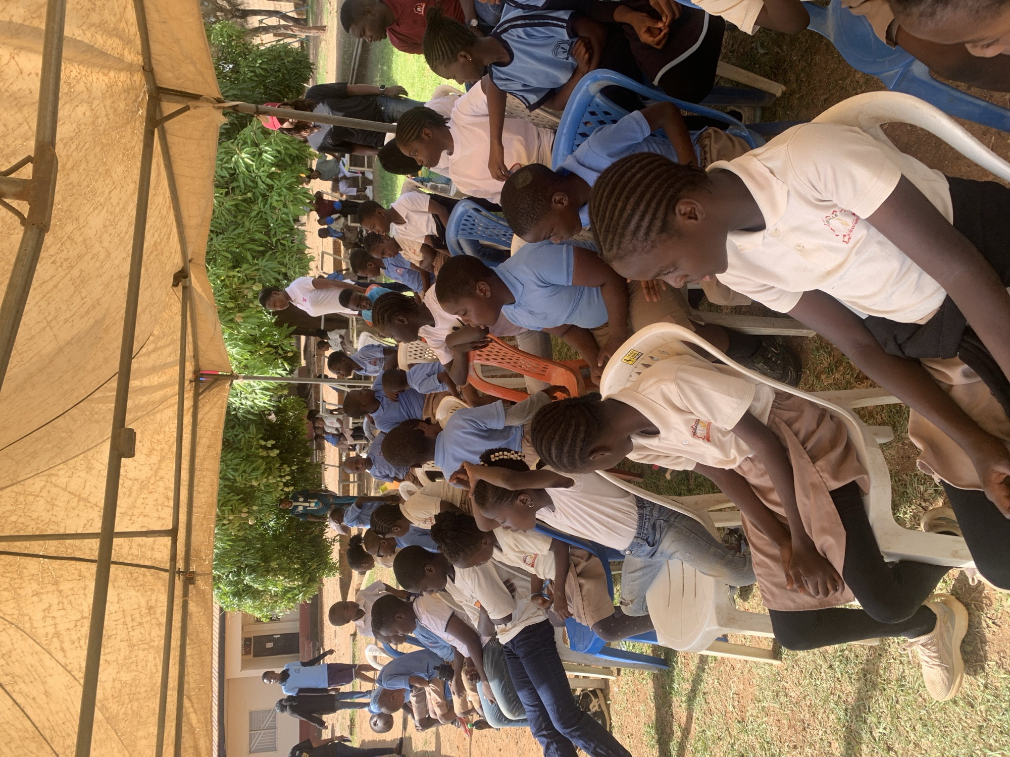 This media file depicts an outdoor gathering under a large, light-colored canvas tent in Jos, Nigeria, likely during the daytime. The setting suggests sunny weather, with the tent providing shade for the participants.
The primary subjects are numerous children and young people, most of whom appear to be students. They are seated in orderly rows on various plastic chairs, including white, blue, and orange ones. Many children wear uniforms: light blue polo shirts or white polo shirts, some featuring a crest-like logo on the chest. Their postures indicate attentiveness, with many looking forward or having their heads slightly bowed, suggesting they are observing or listening intently to an activity or speaker not visible in the frame. A few adults are also present, some standing in the background.
Beyond the seated attendees and the tent structure, lush green foliage, including bushes and trees, is visible. In the background, simple buildings with windows can be seen. The ground is a mix of dirt and sparse grass. One white plastic chair in the foreground has partially visible text on its side, though it is indistinct. The overall scene conveys a sense of community engagement or an educational event.