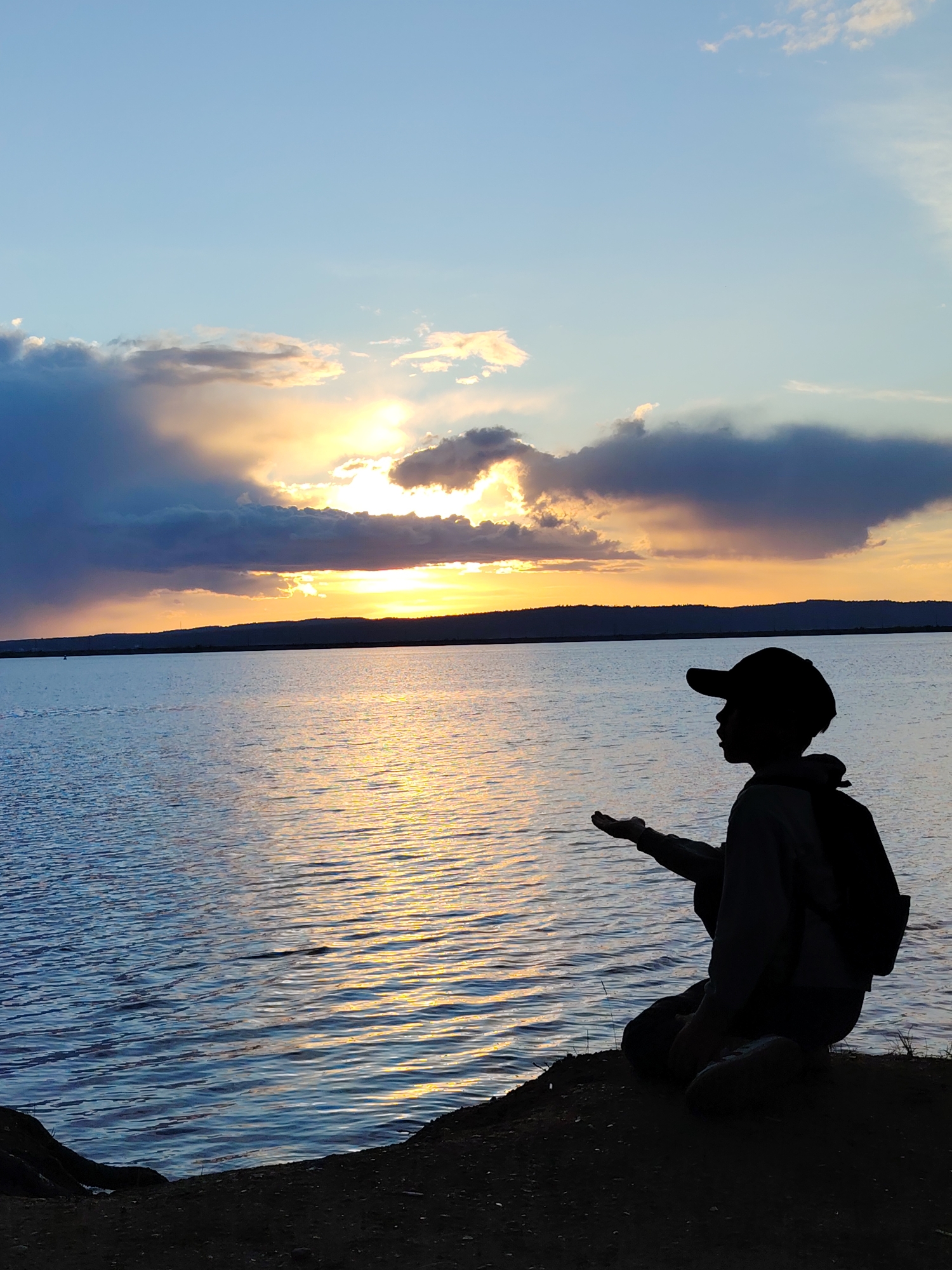 A person wearing a baseball cap and backpack is silhouetted while kneeling by a body of water, facing the sunset. Their hand is outstretched. The background features the sunset over the water and horizon, with cloud formations. The location may be the city of Chaikovsky, Russia, based on the request, but the image does not contain any locational information to confirm that claim.