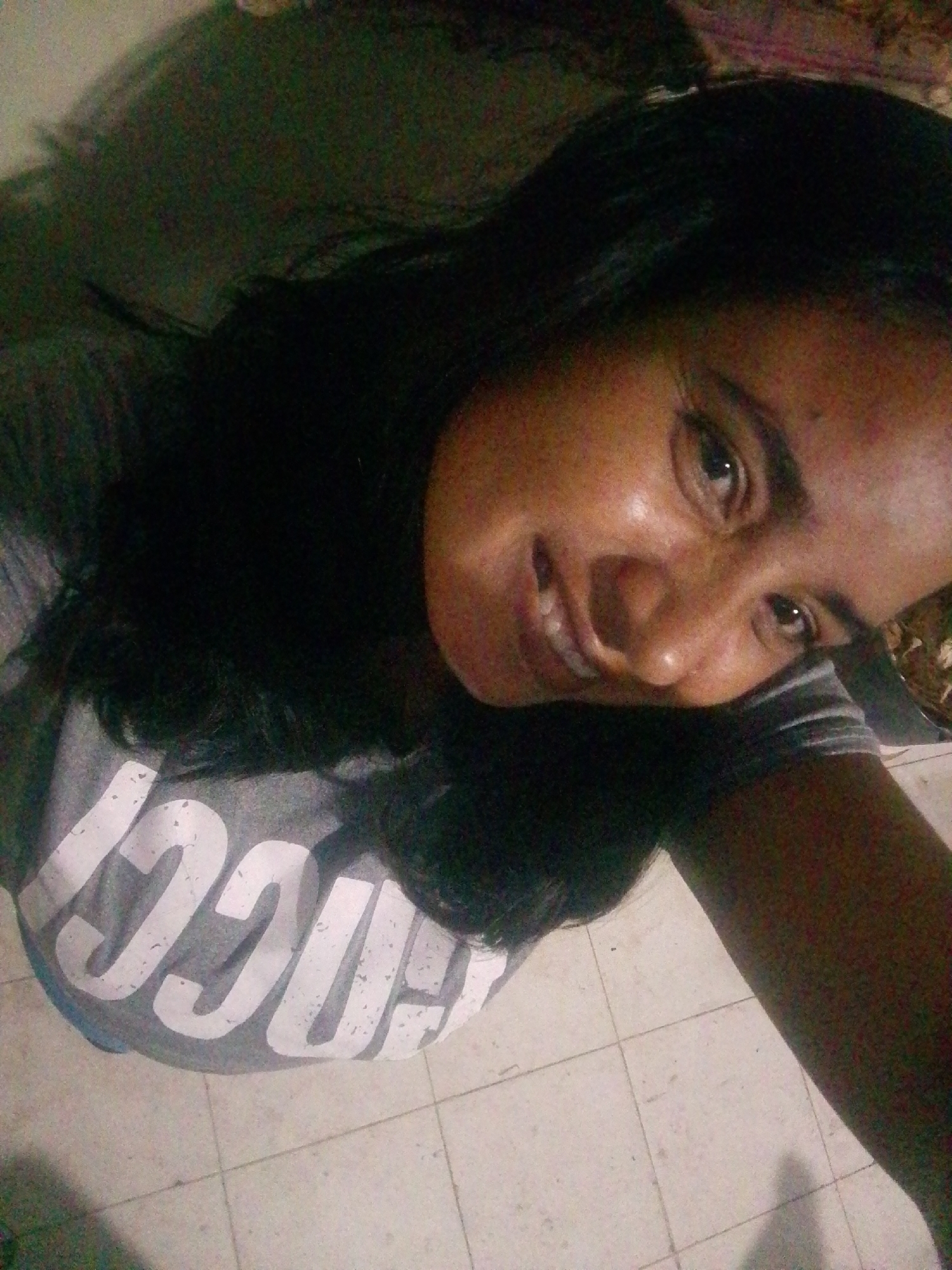 A woman with dark hair and skin smiles at the camera. She wears a grey t-shirt with white lettering. The background includes a light-colored tile floor and indistinct shapes suggesting a room interior. There's no information indicating a location in the city of Juan Griego, Venezuela.