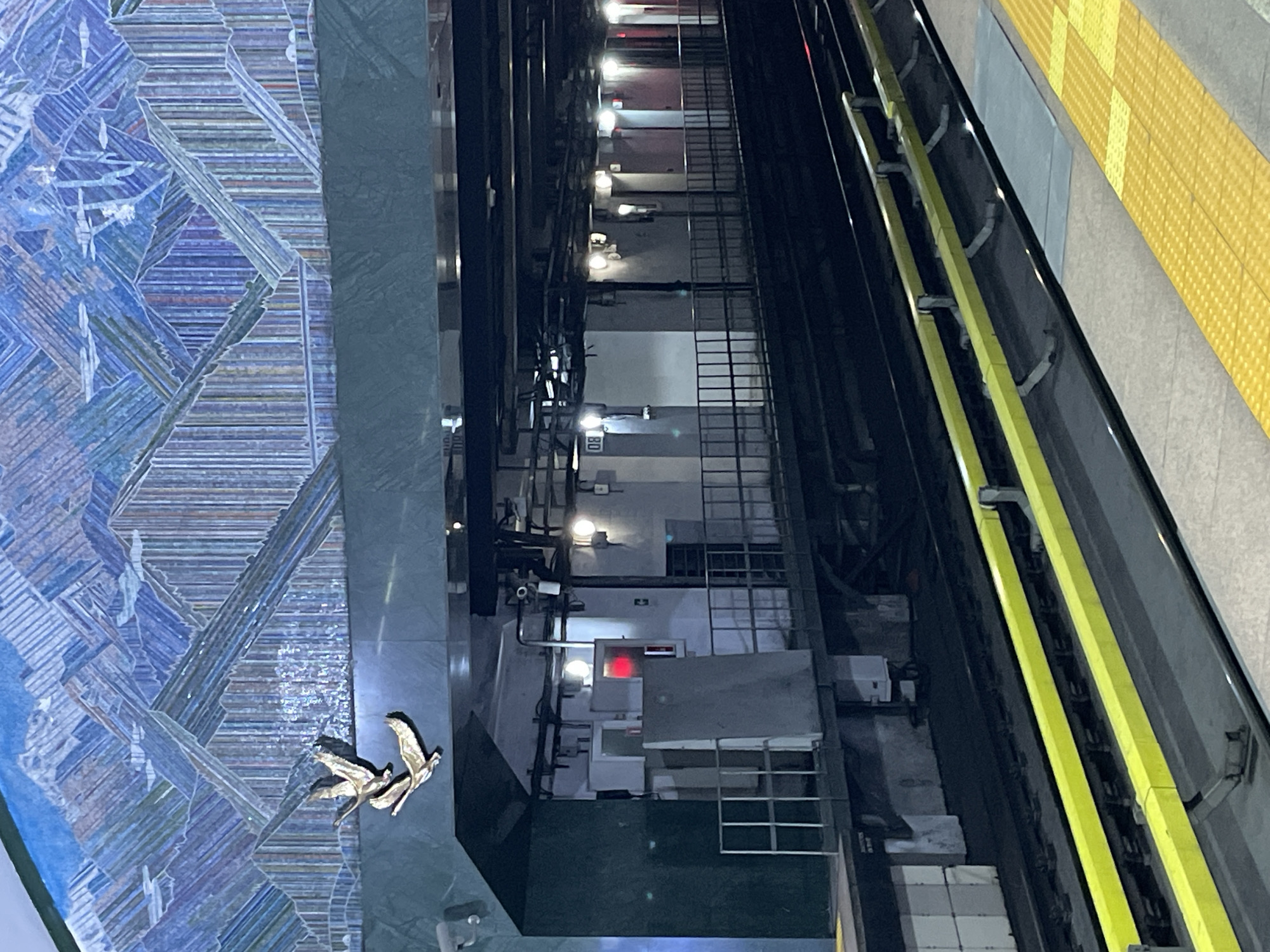 This image shows a view from a subway platform looking down at the tracks and the opposite platform. The scene is a subway station with a mosaic mural on the left wall and yellow tactile paving on the right. Two metallic bird sculptures are placed on the mural.
The train tracks are visible in the center of the image, with overhead wires and lighting. The opposite platform features a series of doors and windows, with some lights illuminating the interior. There are some maintenance equipment and ladders visible. One of the crops shows a red light on a piece of equipment.
The image appears to be taken during the day, but the artificial lighting in the station creates a somewhat dim atmosphere. There is no indication of weather conditions. The location is a subway station, as evidenced by the tracks, platform, and signage. The overall mood is neutral, with no discernible emotions.
Visible text includes "80" on a sign in the distance.