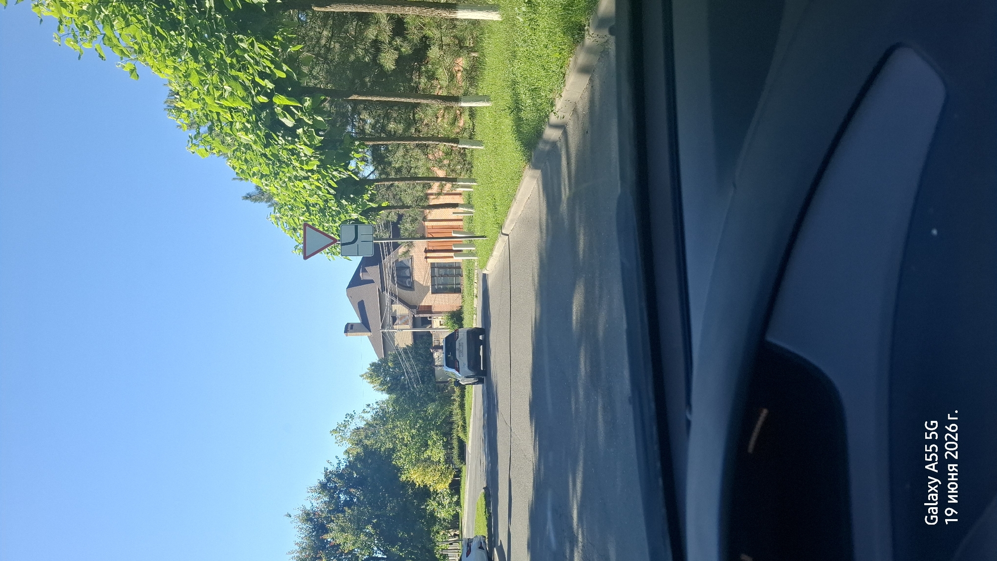 This is a vertical, full-shot, eye-level photograph taken from inside a vehicle, looking out at a residential street. The scene depicts a sunny day with a clear blue sky. The foreground is dominated by the dark blue interior of the car, specifically the door panel and window frame, which obscures a significant portion of the view.
The main subjects are a street lined with green trees and a few houses. On the left side of the street, there are tall, leafy trees casting shadows. Beyond the trees, a two-story brick house with a dark roof and multiple windows is visible. A road sign, a triangle with a red border and an arrow indicating a turn, is positioned above the house. Further down the street, another white car is parked. The street itself is paved and appears to be in good condition. The right side of the street features a well-maintained grassy verge and more trees.
The timestamp in the bottom right corner indicates "Galaxy A55 5G 19 июня 2026 г." which translates to "Galaxy A55 5G June 19, 2026." This suggests the image was captured on that date. The bright sunlight and clear sky indicate it's daytime, likely late morning or early afternoon. The overall mood is peaceful and mundane, typical of a suburban street scene. There are no people visible in the photograph. The location context provided is Rostov-on-Don, Russia, which aligns with the architectural style of the house and the presence of deciduous trees common in the region.