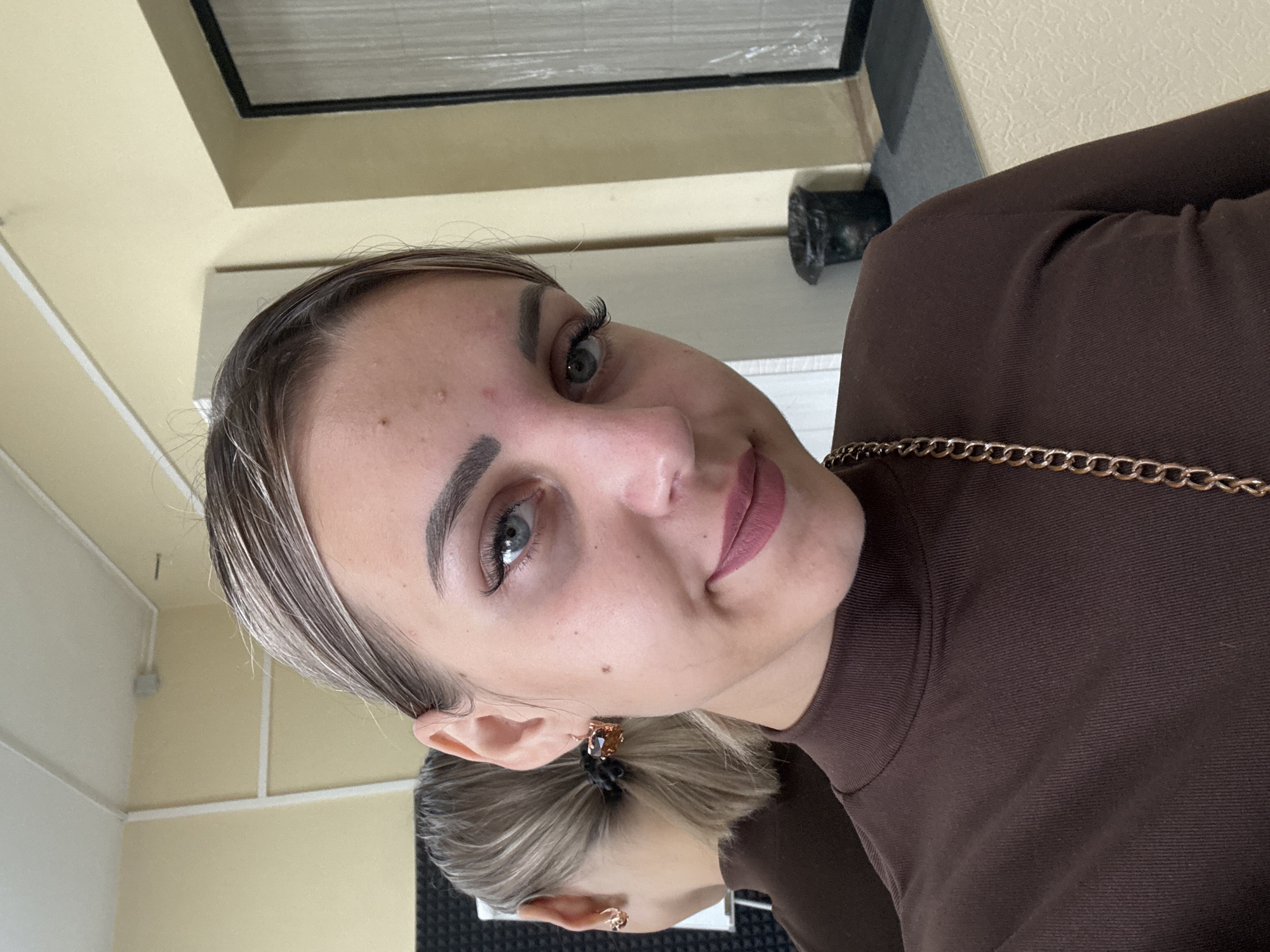 A young Caucasian woman is shown in a close-up selfie, her head and shoulders filling the frame. She has light skin with some minor blemishes on her forehead and cheeks. Her eyebrows are dark and well-defined, and she is wearing subtle eye makeup, including false eyelashes. Her hair is pulled back into a ponytail, with some lighter streaks visible. She is wearing a dark brown, high-necked top, and a gold chain is visible across her chest. She has large, square-shaped earrings with a bronzed crystal. Her left ear is visible, and a reflection of her right side, including her ponytail, is seen in a mirror to her left. The background appears to be an interior space with light yellow walls and white trim. A black trash can is visible in the background on the right side. The lighting is moderate, and the image is slightly grainy. The overall impression is that of a casual selfie taken indoors, possibly in a dressing room or personal space. There are no specific landmarks or details that definitively place the image in Yuzhno-Sakhalinsk, Russia.