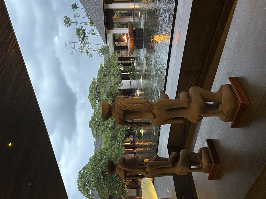 The image captures an outdoor area of a resort or hotel in Nusa Dua, Indonesia, under a heavily overcast, grey sky. In the foreground, two dark-colored statues of female figures, sculpted from stone or wood, are positioned on dark wooden bases on a dark tiled floor. Both statues are seated, facing away from the viewer, with braided hair designs and bowls atop their heads. They are situated alongside a dark, low retaining wall separating the foreground from a large, dark water feature. This water feature contains multiple illuminated fountains, creating visible ripples on its surface, and centrally features a large, dark bowl holding an active open flame. Beyond the water, traditional Balinese-style architectural elements with dark, tiled roofs and supporting pillars are visible, interspersed among dense green trees and taller, slender trees in the background. The scene is devoid of any individuals.