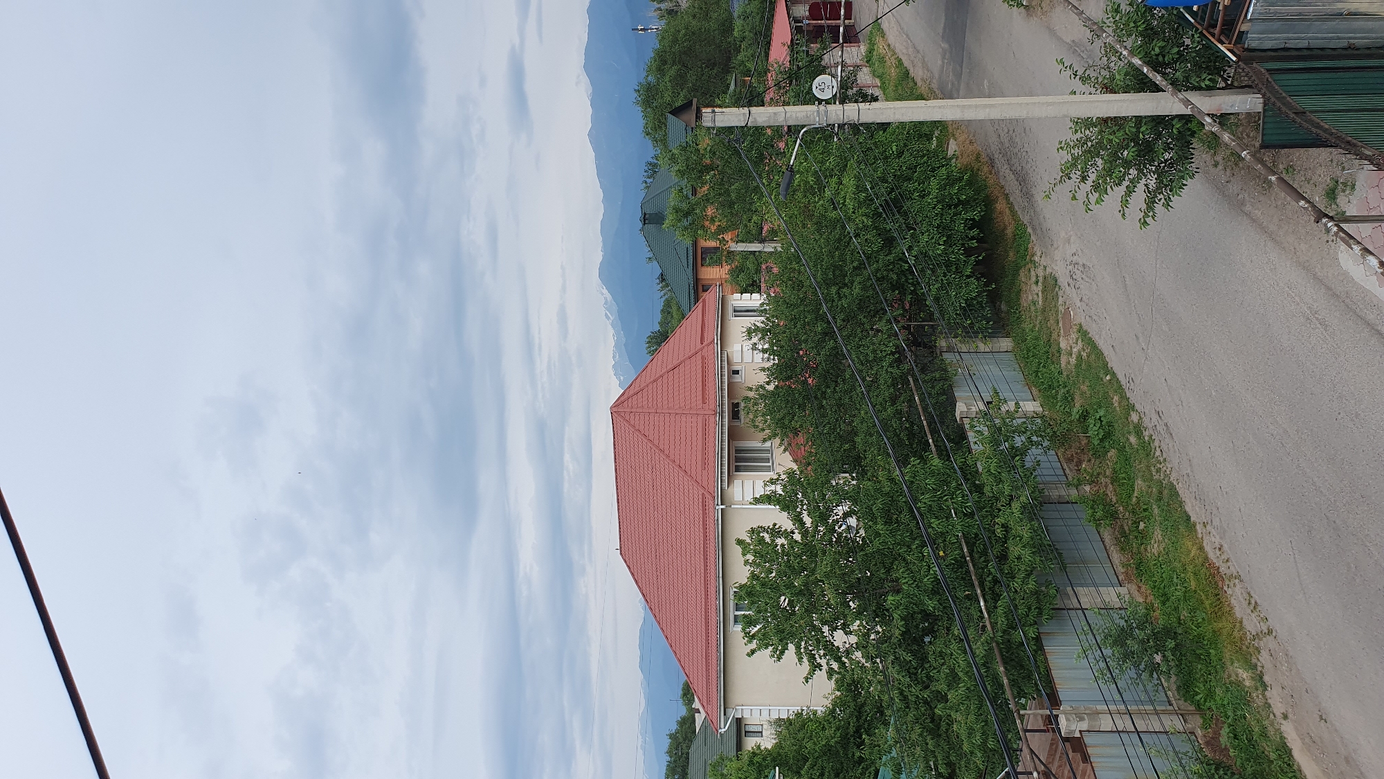 This image depicts a residential area in Almaty, Kazakhstan, likely during daytime given the bright sky. The scene is dominated by a two-story house with a prominent red, pyramid-shaped roof. The house is light-colored and has several windows. It is surrounded by lush green trees, some of which partially obscure the building. In the background, more houses with different colored roofs are visible, nestled amongst trees. Further in the distance, imposing mountains with hints of snow on their peaks rise against a pale blue sky filled with wispy clouds.
A paved road curves through the scene, bordered by a metal fence and more greenery. A concrete utility pole stands near the road, supporting electrical wires that crisscross the image. One of the utility poles has a circular sign with a white background and black numerals, possibly indicating a street number or route, though the text is not fully legible. The overall impression is one of a quiet, suburban or semi-rural setting, possibly in the foothills of the mountains that are characteristic of Almaty's landscape. There are no people visible in the image. The weather appears to be clear with some cloud cover, suggesting a pleasant day.