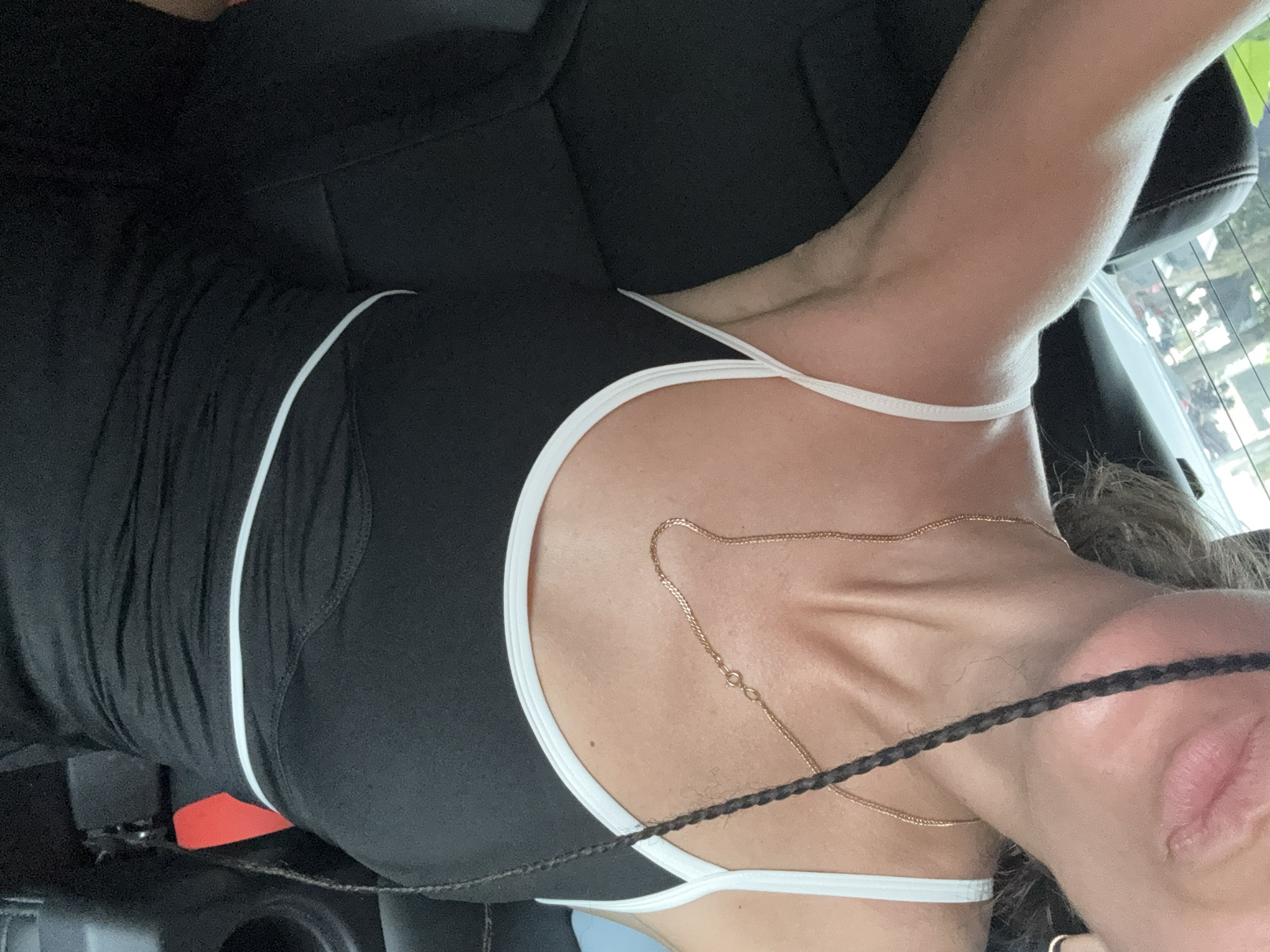 The image shows a close-up of a woman in the backseat of a car. She is wearing a black tank top with white trim and a gold necklace. Her arm is raised, and a braid of her hair hangs down her chest. The background is blurred, showing trees and buildings outside the car window. There is no specific indication of the location being Hoa Lư Ward, Vietnam.
