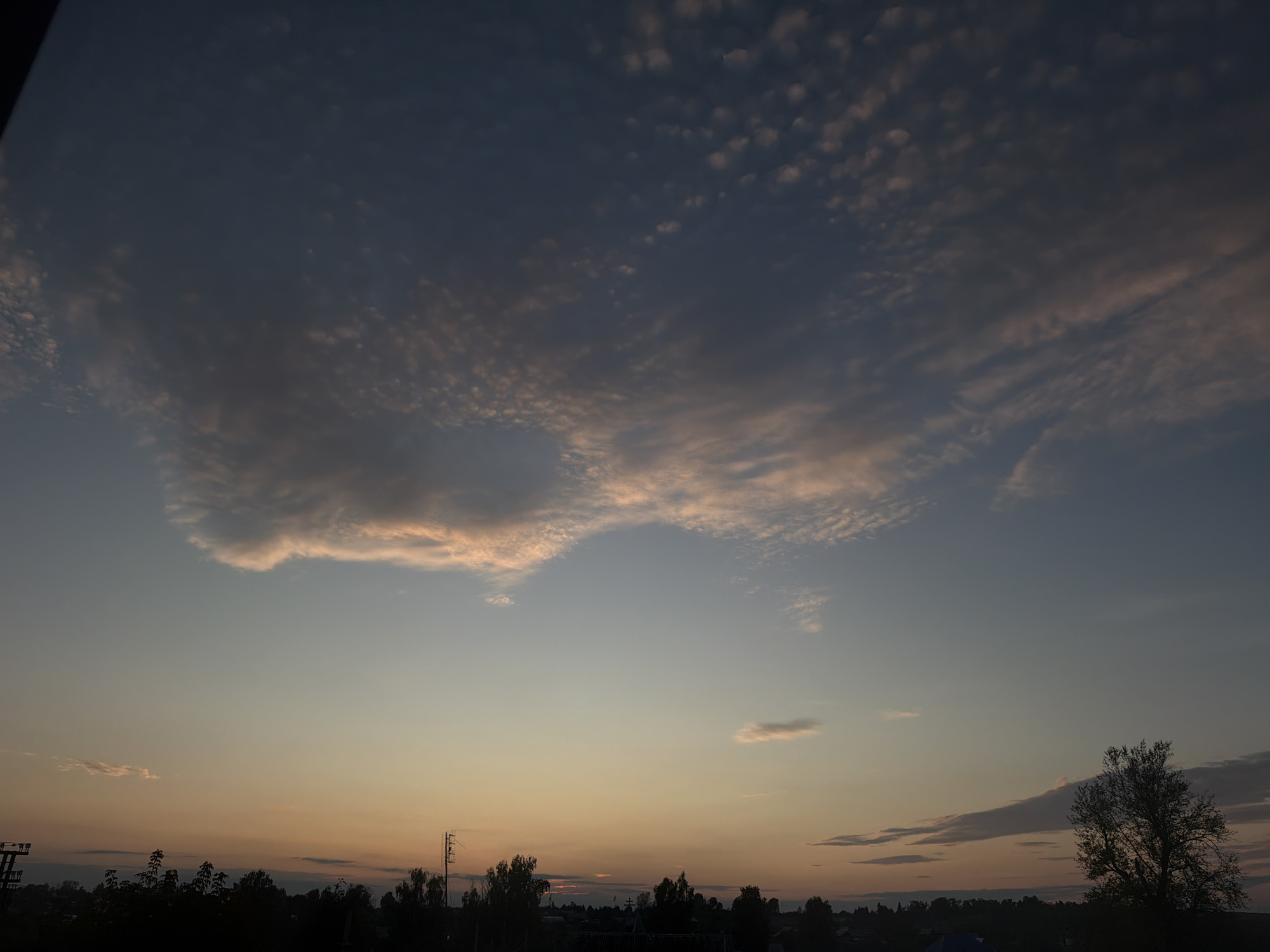 A wide-angle, eye-level shot captures a twilight sky over a dark, silhouetted landscape. The sky is a gradient of colors, transitioning from a deep blue at the top to a pale orange and yellow near the horizon. Wispy clouds, tinged with pink and orange, are scattered across the upper portion of the sky, with a larger, more textured cloud formation dominating the center. Below the sky, a dark band of trees and buildings forms the horizon line. In the foreground, the silhouettes of trees are prominent, with a single, larger tree on the right side of the frame. Power lines and poles are visible on the left and right sides of the image. The scene depicts a rural or suburban setting at dusk, with no people or animals visible.