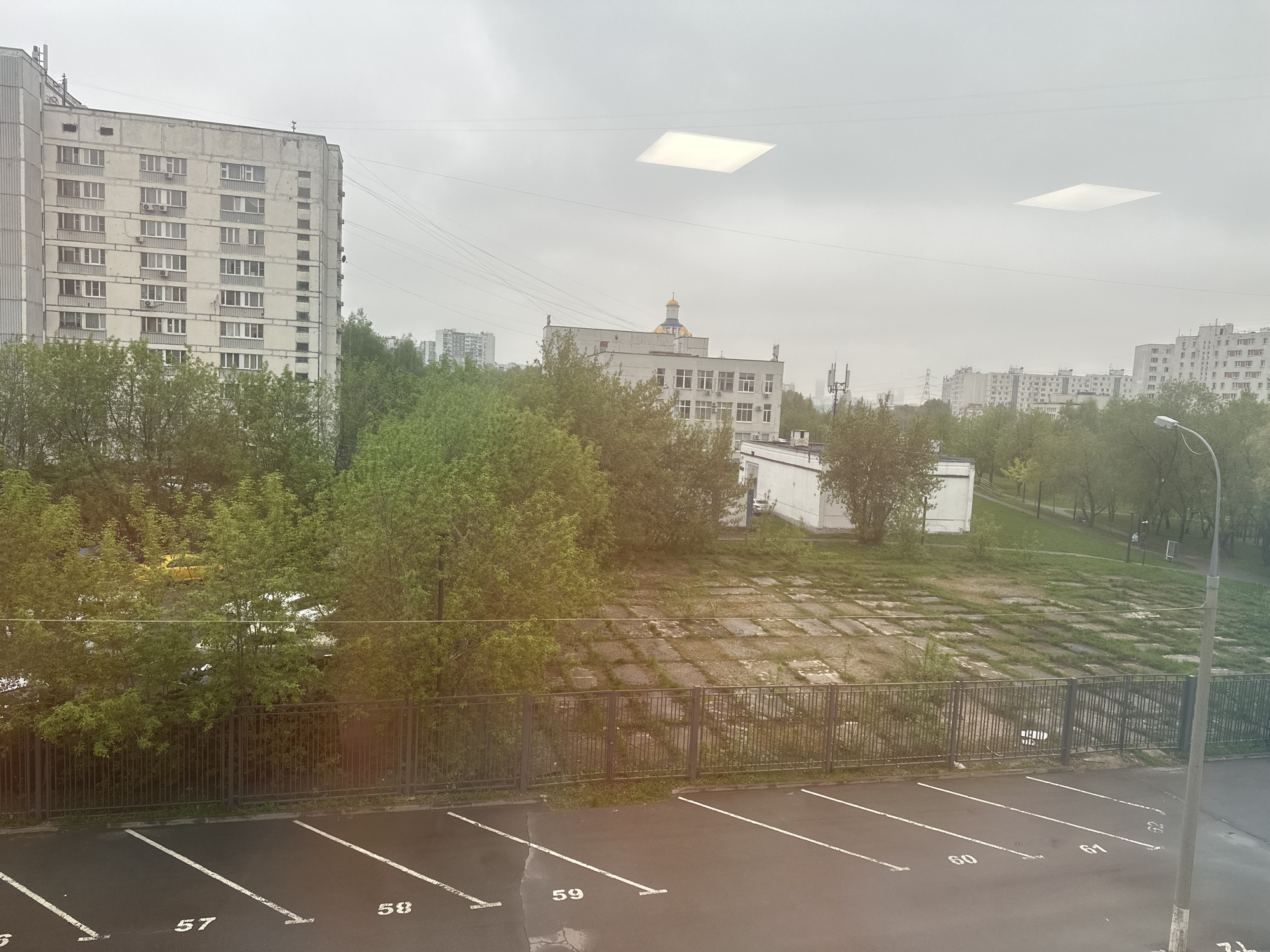 The image depicts a view from a window in Moscow, Russia. In the foreground is a wet, black asphalt parking lot marked with white parking space lines numbered 57 to 61. Beyond the parking lot is a low metal fence separating it from a grassy area covered in rectangular concrete slabs with vegetation growing in the gaps. In the middle ground are several deciduous trees with green leaves. Behind the trees is a white building with a gold dome visible above the roof, likely a church. To the left is a tall, grey apartment building. Other apartment buildings are visible in the distance on the right. The sky is overcast and grey, with two bright square reflections visible on the window, presumably from interior lighting. A streetlight is positioned in the right foreground. The environment appears urban.