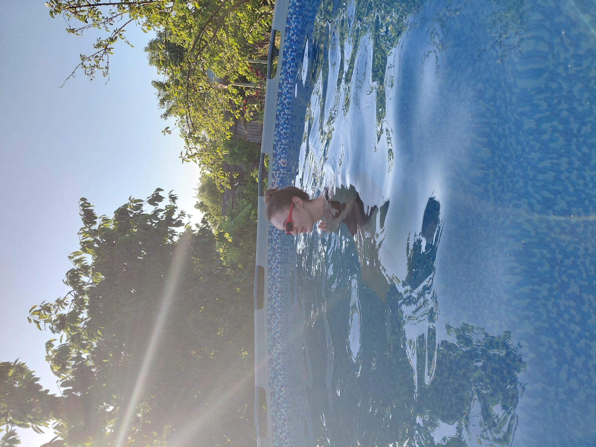 In this image, a person wearing red sunglasses is submerged in a swimming pool. The water is blue and reflects the surrounding trees and sky. The person's head is tilted slightly, and they appear to be looking down. The pool has a patterned border with blue and white tiles. Sunlight streams in from the left, creating lens flare and illuminating the water and surrounding greenery. The background shows lush green trees and a clear blue sky, suggesting it is a sunny day. The overall impression is one of relaxation and enjoyment on a warm day. The image is oriented vertically, with the top of the frame showing the sky and trees, and the bottom featuring the pool and the person.