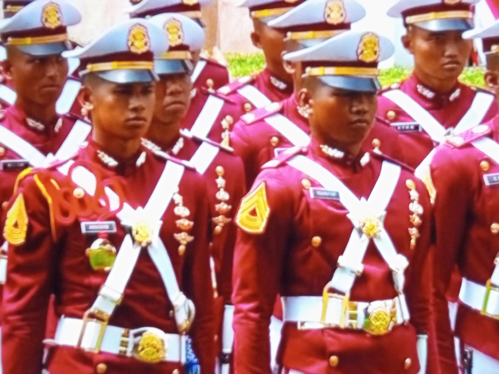 The image shows a group of young men in uniform marching in what appears to be a parade or formal event. They are wearing distinctive maroon jackets with gold trim, white cross-body straps, and white belts with ornate buckles. Their caps are light blue with gold insignia. The men are all facing forward and appear to be in formation. The background is slightly blurred but suggests an outdoor setting with some greenery and possibly buildings. The detail on their uniforms, including medals and badges, indicates a military or cadet setting. The specific location in Banyudono, Indonesia, is not directly evident from the visual information in the image itself but is provided as context.