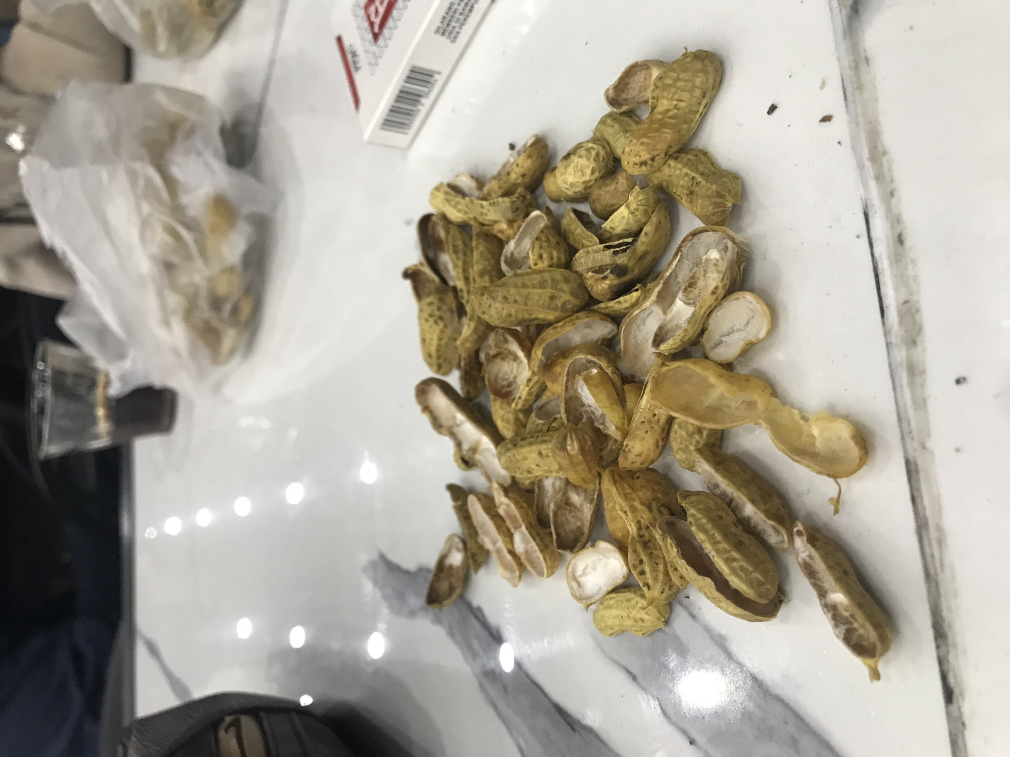 The image shows a pile of empty peanut shells scattered on a white surface with a marble-like pattern. Next to the shells is a partially transparent plastic bag containing what appears to be more peanuts, and a package of cigarettes. The photograph appears to be taken indoors. No people are visible. The scene is presented on a table that is possibly made of a synthetic surface. There is no way to determine the location is in Lhokseumawe, Indonesia.
