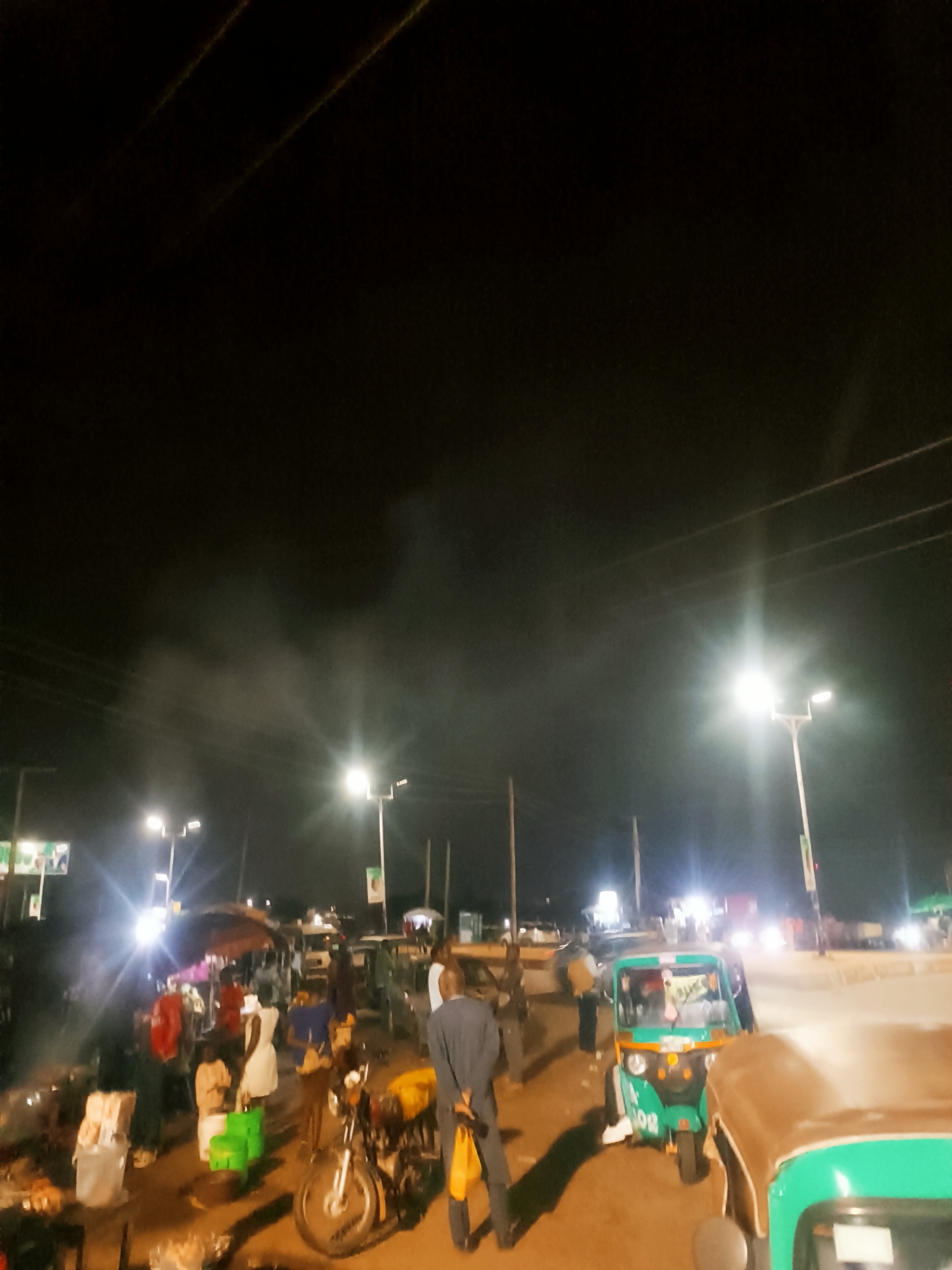 The image depicts a bustling street scene at night in Bukuru, Nigeria. The foreground features a motorcycle parked near a man in a suit holding a yellow bag. Several green three-wheeled vehicles, or tuk-tuks, are also visible. In the middle ground, a small market is illuminated by streetlights and appears to be active with people and stalls. Overhead, utility lines are visible against the night sky, which is partially obscured by what appears to be smoke or mist near the lights.