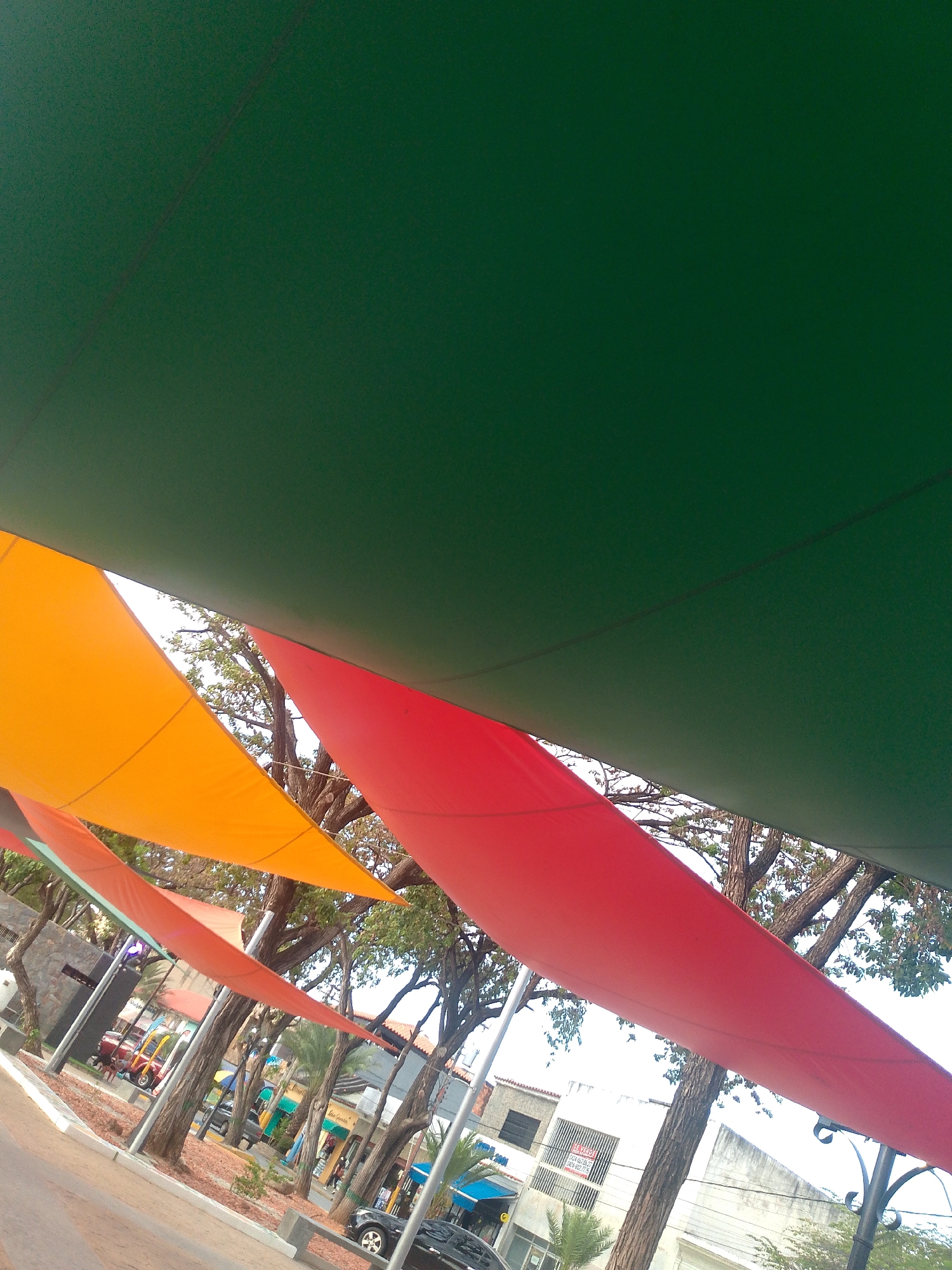 The image depicts an outdoor scene in Juan Griego, Venezuela, featuring colorful shade sails stretched overhead. The sails are green, red, and orange. Below the sails, a street scene is visible with trees, buildings, parked cars, and pedestrians. The angle of the shot is upward, emphasizing the colorful sails and the urban environment beneath.