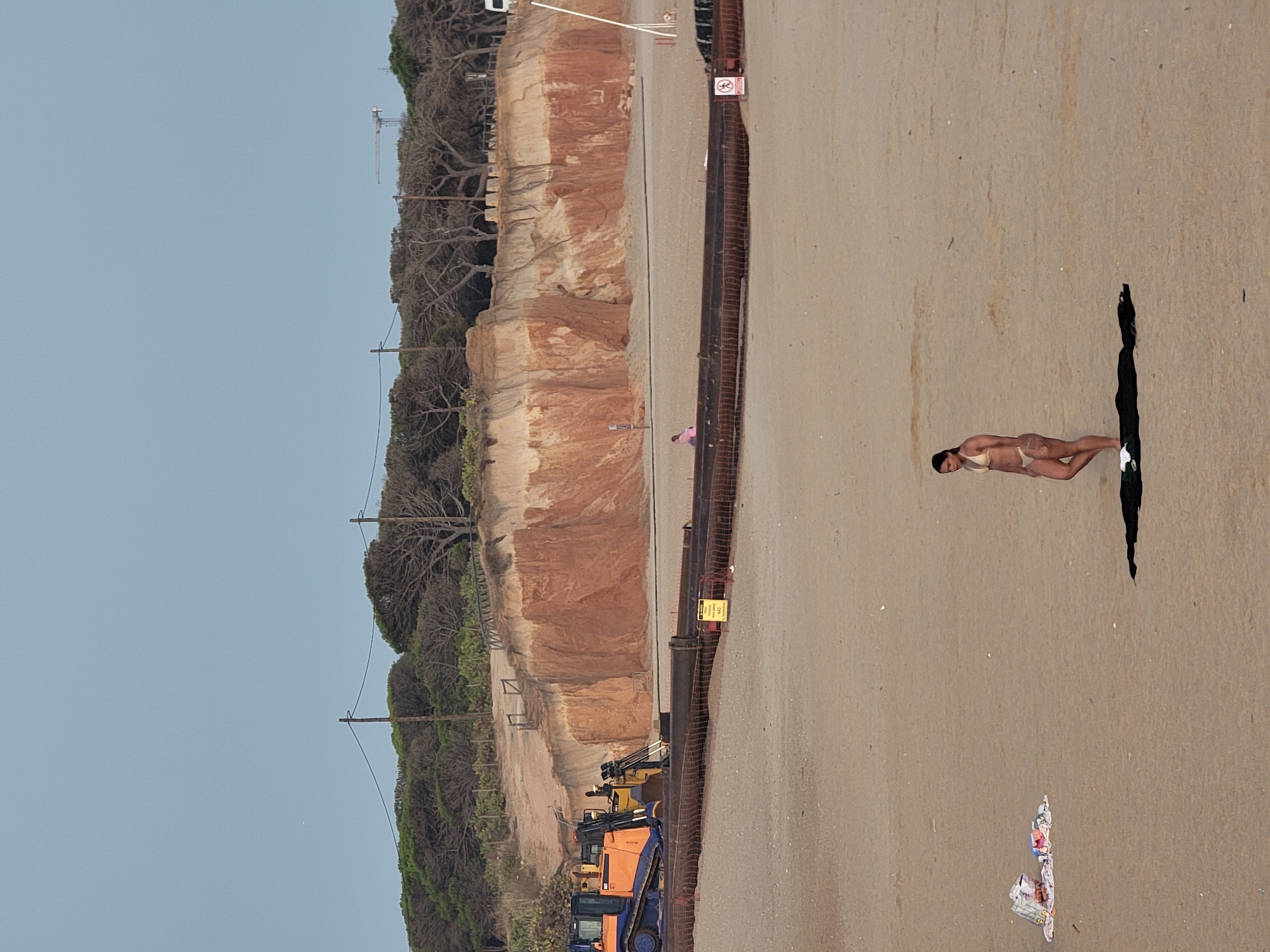 The image depicts a beach scene with a woman in a bikini standing on a black cloth in the foreground. To her left, a colorful towel and other items are scattered on the sand. In the middle ground, construction equipment, including a bulldozer, is visible near a cliff face. Pipes and netting suggest ongoing construction or shore protection work. The background shows a tree-lined cliff overlooking the beach. The scene appears to be located in Quarteira, Portugal, given the context.
