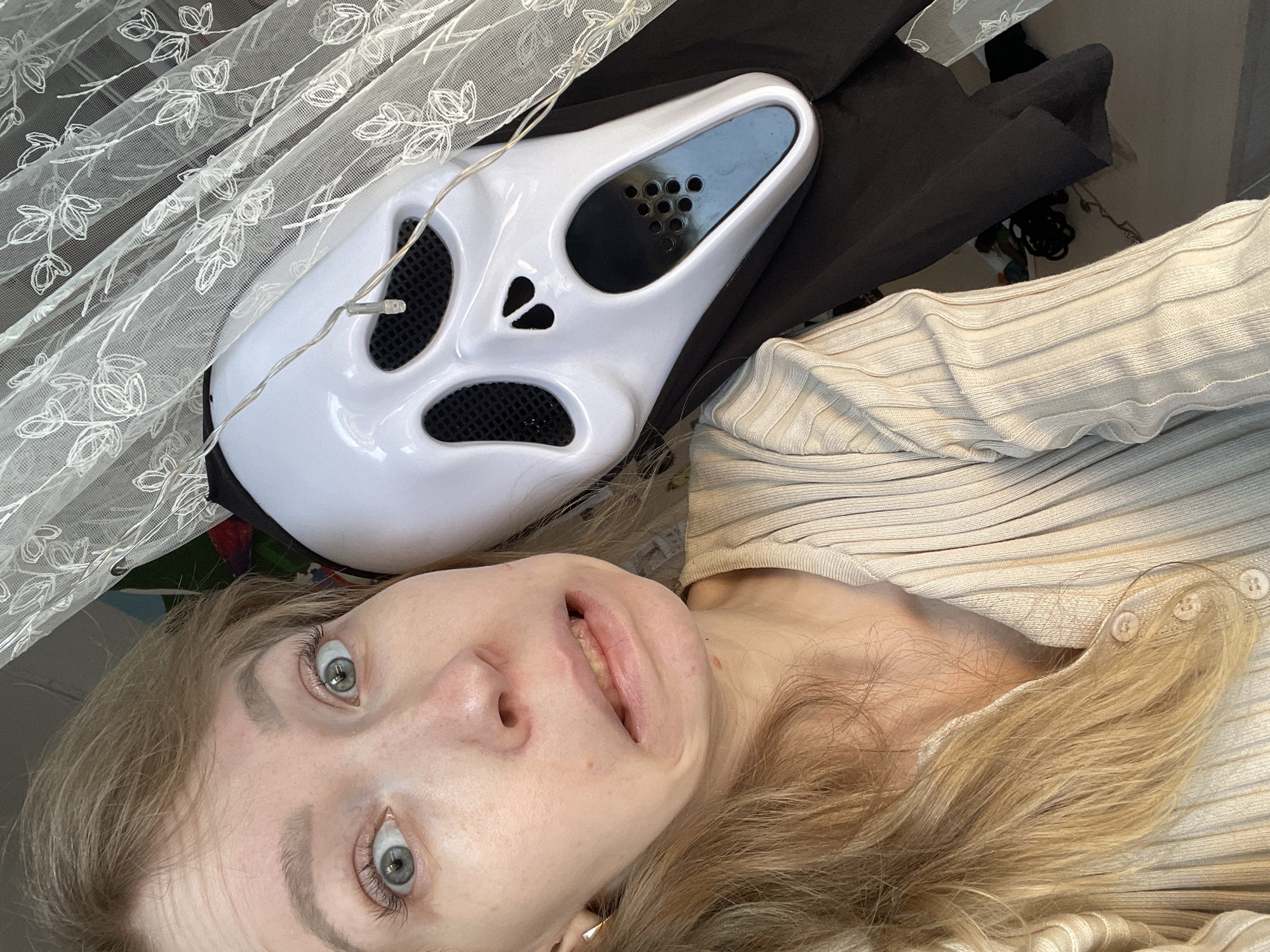 A selfie-style photograph depicts a fair-skinned person with blonde hair, lying down, with a Scream mask placed near their head. The mask is white with black mesh over the eye and mouth openings and is positioned to the left of the person's face. The person is wearing a beige ribbed sweater. A white, floral-patterned lace curtain hangs in the background. A string of small lights is draped near the mask. The location is unknown and cannot be determined based on the image. The city of Siva, Russia, is irrelevant as the photo's location is not discernible.