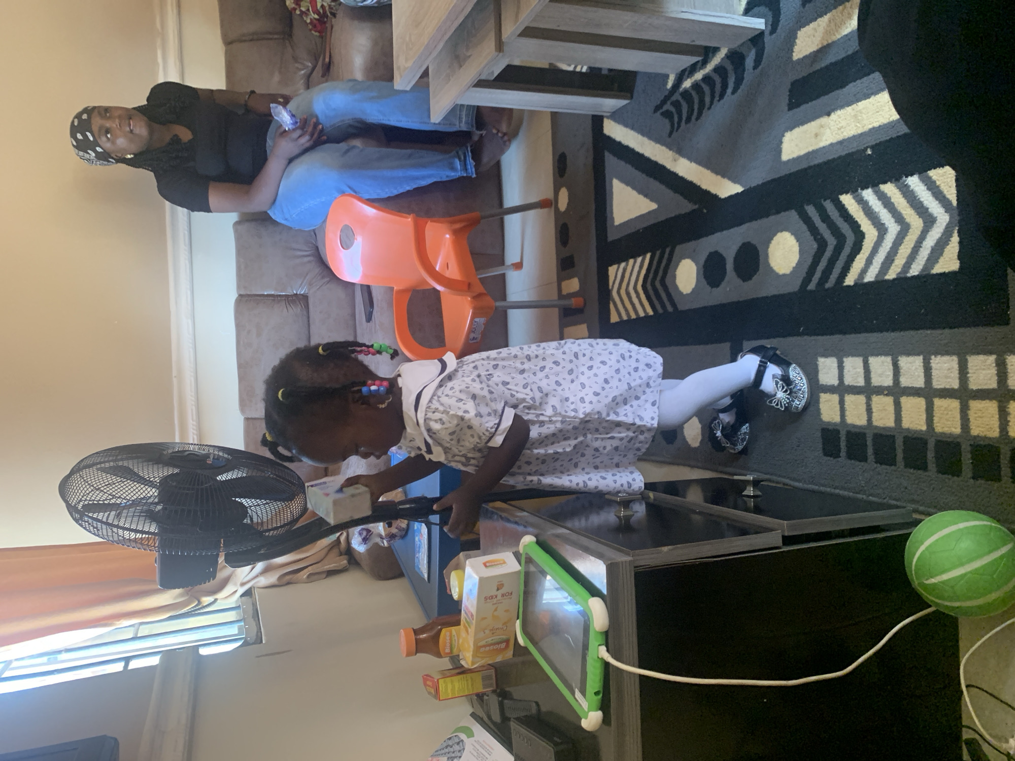In Gura Top, Nigeria, a young child stands in front of a black oscillating fan, holding a white box. The child is wearing a white dress with a patterned print and white tights, along with embellished shoes. Nearby on a dark wooden table, a tablet with a green case is plugged in and charging, alongside several juice boxes. To the right, a woman with braided hair is seated on a brown couch, wearing a black t-shirt and blue jeans, and a black and white bandana. An orange plastic chair is positioned between the child and the woman. The room has light-colored walls, a patterned rug on the floor, and a wooden table to the right. Sunlight streams in from a window covered by peach-colored curtains.