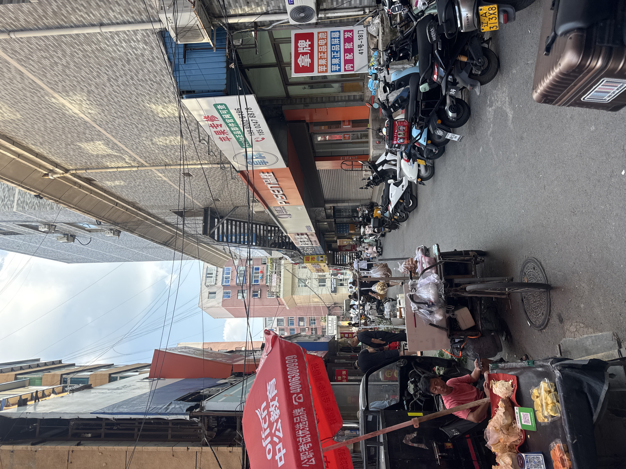 The image depicts a narrow street scene in Dadong District, China, likely during the daytime. Buildings line both sides of the street, featuring a mix of brick and concrete facades, with some showing signs of wear and tear. Electrical wires crisscross overhead.
On the left side of the street, a vibrant red umbrella advertising "offich" and "中公教育" shades a food stall. A man in a pink shirt is seated behind a table laden with produce, including what appear to be dried fruits and root vegetables. A black tricycle-like vehicle is parked nearby.
The right side of the street is occupied by parked scooters and motorcycles. Several shops are visible, including one with a sign advertising "苹果专修" (Apple Repair) and another with a red sign that reads "拿牌" (Take a card/Get a card), which also lists "苹果正品电池" (Genuine Apple Battery) and "苹果正品屏幕" (Genuine Apple Screen). A blue metal structure is also present.
People are scattered throughout the scene, some standing and conversing, others walking or tending to their stalls. The overall impression is of a busy, active urban environment with elements of commerce and daily life.