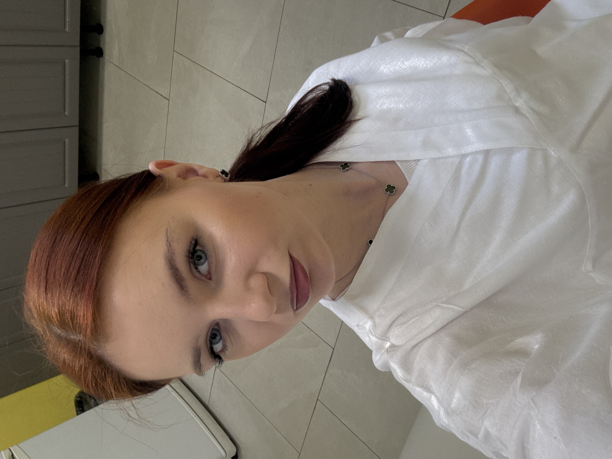 A woman with auburn hair and fair skin is shown from the chest up, wearing a white shirt and necklace. She appears to be indoors, possibly in a kitchen based on the cabinets and countertop partially visible in the background. The location in the city is Unknown, Unknown.