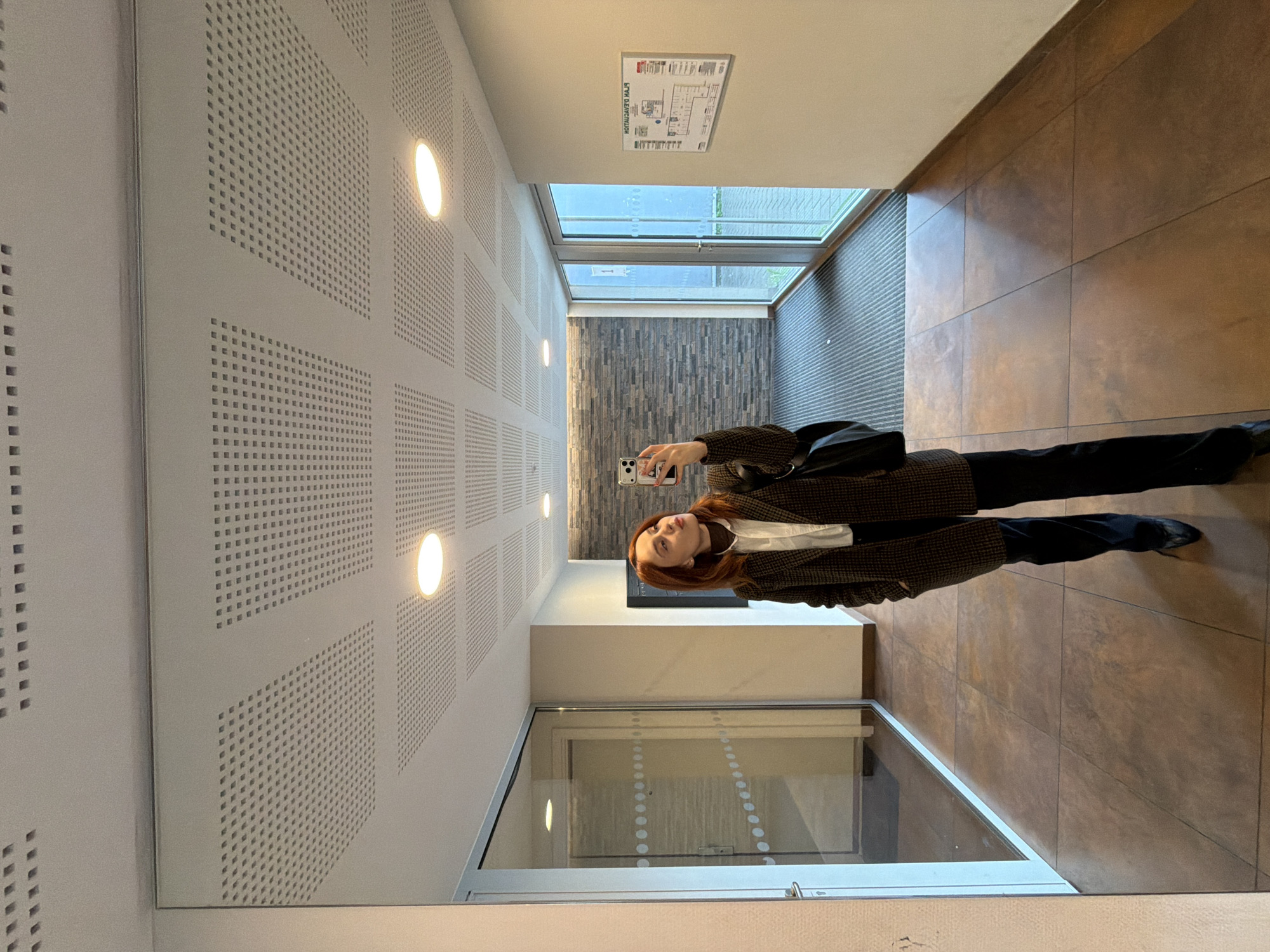 A woman with reddish-brown hair, dressed in a white collared shirt, a dark brown plaid blazer, black trousers, and black pointed-toe shoes, is reflected in a large, floor-to-ceiling mirror within a modern hallway. She holds a white smartphone with multiple cameras up to her face, capturing a selfie, with her gaze directed towards the phone's lens. A black shoulder bag is visible on her left shoulder.
The environment is a well-lit interior corridor, characteristic of a contemporary institutional or commercial building in Strasbourg, France. The floor is covered with large, reddish-brown square tiles, transitioning to a grey ribbed mat near the far end. The right wall is off-white, featuring a rectangular "PLAN D'ÉVACUATION" (Evacuation Plan) sign, reflected reversed as "LÀJ NOITAUCAVE NALP". The wall at the end of the hallway is clad with horizontal grey stone-like panels. The ceiling consists of white acoustical tiles with a grid of small square perforations, equipped with multiple circular recessed light fixtures. Large glass double doors with metal frames, marked with a vertical white number "1" on the left pane, are visible at the hallway's end, looking out onto an exterior paved area. A further glass door with horizontal dot patterns is visible to the left in the mirror's reflection.