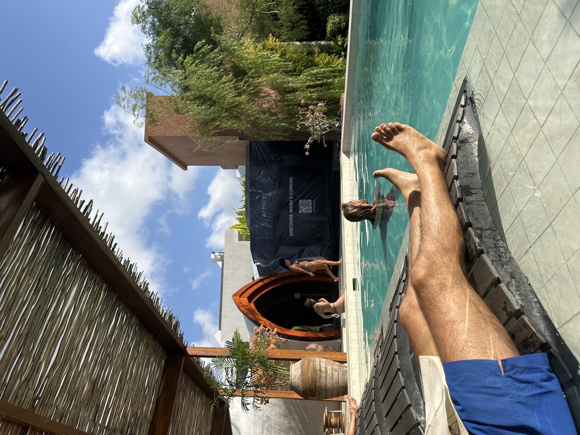 In Kuta Selatan, Indonesia, on a bright, sunny day, a person relaxes on a lounger by a swimming pool. The lounger is made of dark wood planks with a textured surface and is partially submerged at the edge of the pool. The person's legs, from the knees down, are visible, wearing blue swim shorts. Their bare feet are propped up on the lounger, facing the pool. The pool water is clear and turquoise.
In the background, a woman in a bikini walks along the pool edge, emerging from a large, arched wooden doorway. Another woman is sitting on the edge of the pool, also in a bikini. Further back, a black banner with white text that reads "ATMOS DISCOVER WHAT'S COMING" is displayed. To the right of the banner, lush green trees with weeping branches cascade down, and a tall, terracotta-colored building is visible. Overhead, a thatched roof structure provides shade over the area, with wooden beams and natural branches forming the canopy. The sky is a clear blue with scattered white clouds.