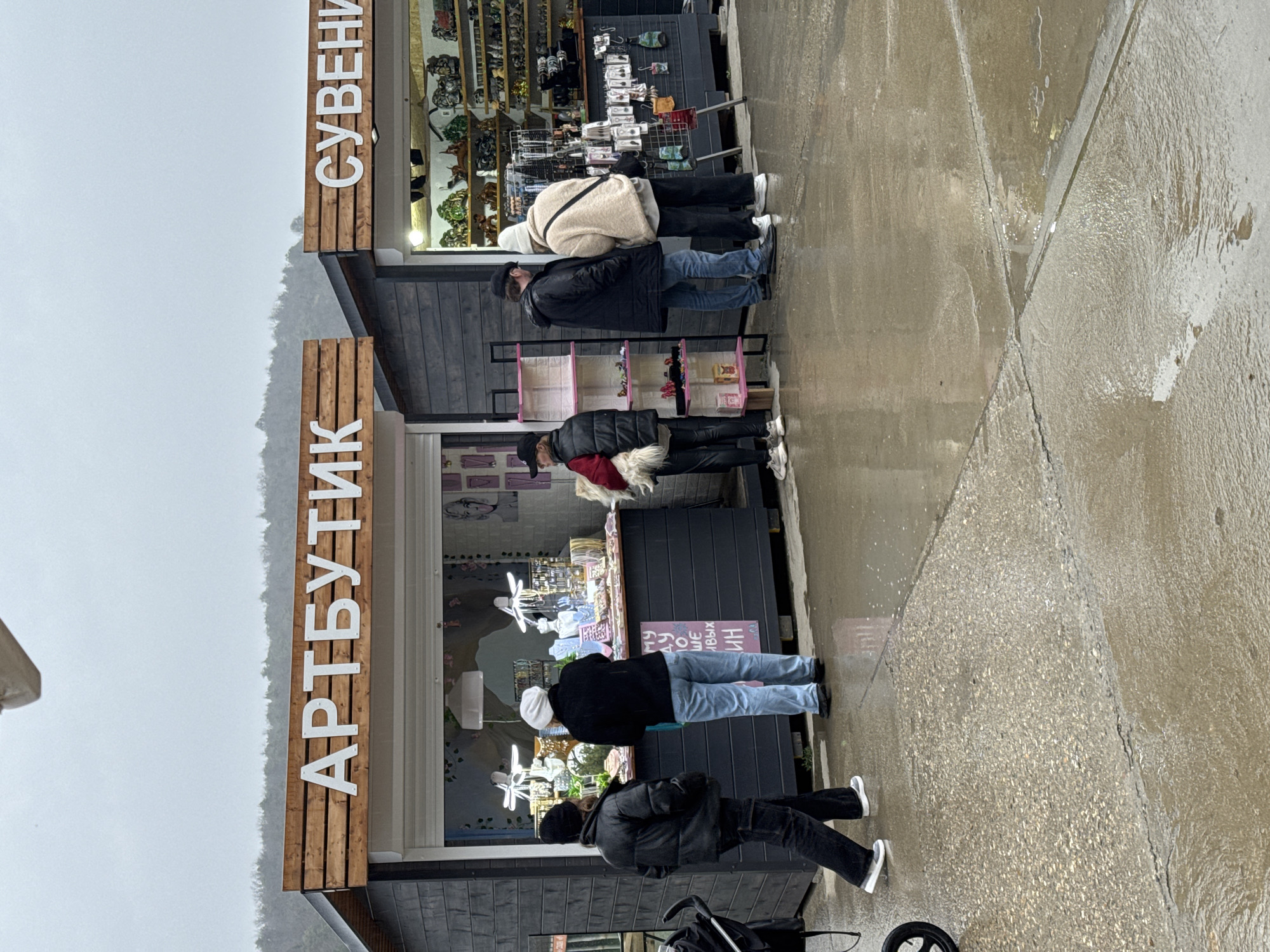 The image shows a street scene in Sukko, Russia. There are two small shops with signs written in Cyrillic, "АРТБУТИК" (Art Boutique) and "СУВЕНИ" (Souvenirs). Five people are looking at the shop displays, standing close to each other on a wet, light-colored, pavement. The shops have dark gray facades, and the signage is white on wood. The sky is overcast and gray. A black baby stroller is partially visible in the lower right corner. The shops appear to be located in a commercial area.