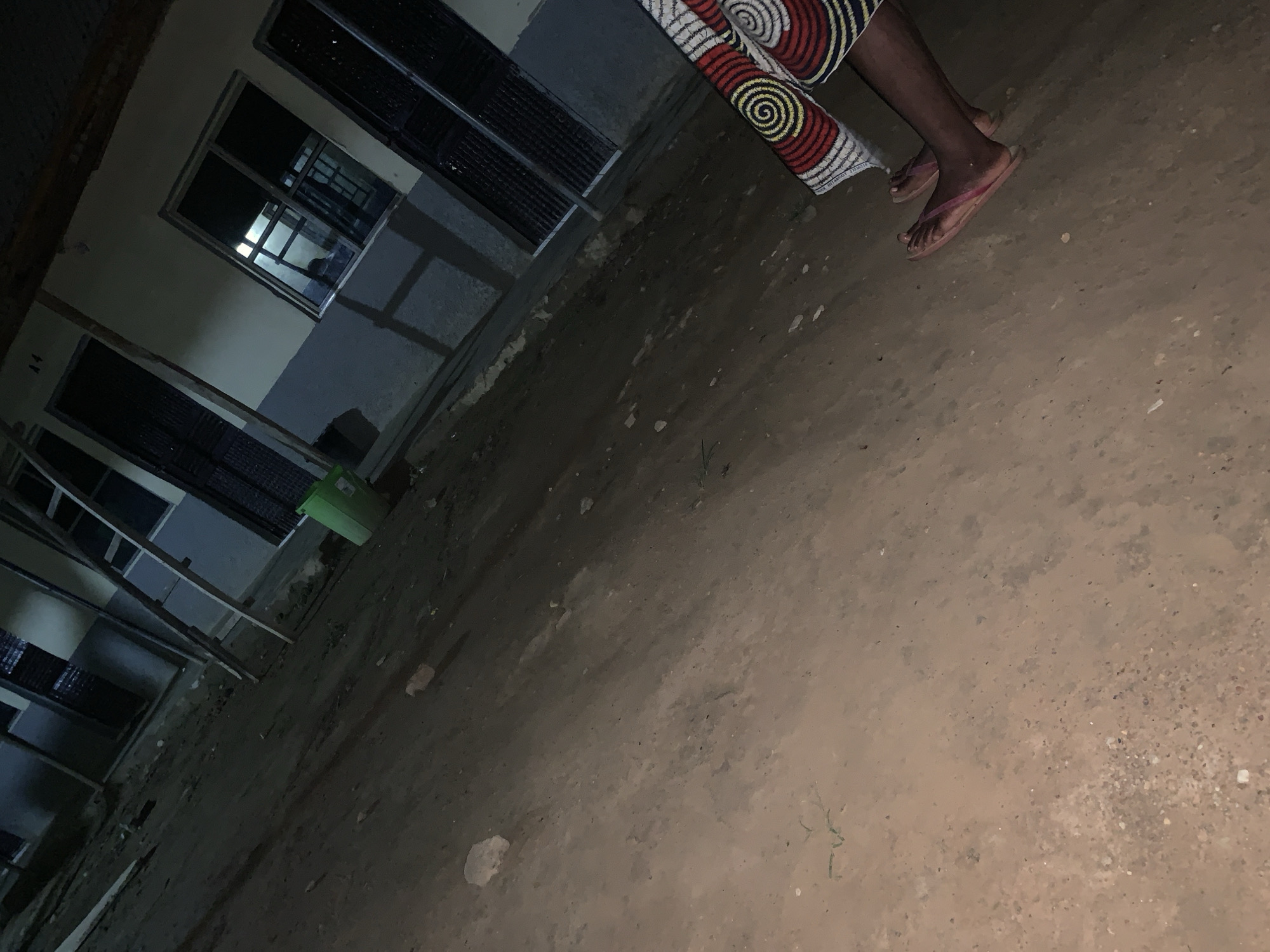 The scene depicts an exterior shot in Keffi, Nigeria, at night. A person wearing a patterned fabric, likely a skirt or dress, and pink sandals stands on a dirt or sandy ground. A building is visible in the background featuring barred windows and a two-tone paint scheme on the walls. There is also a green bucket near the building. The lighting is dim.