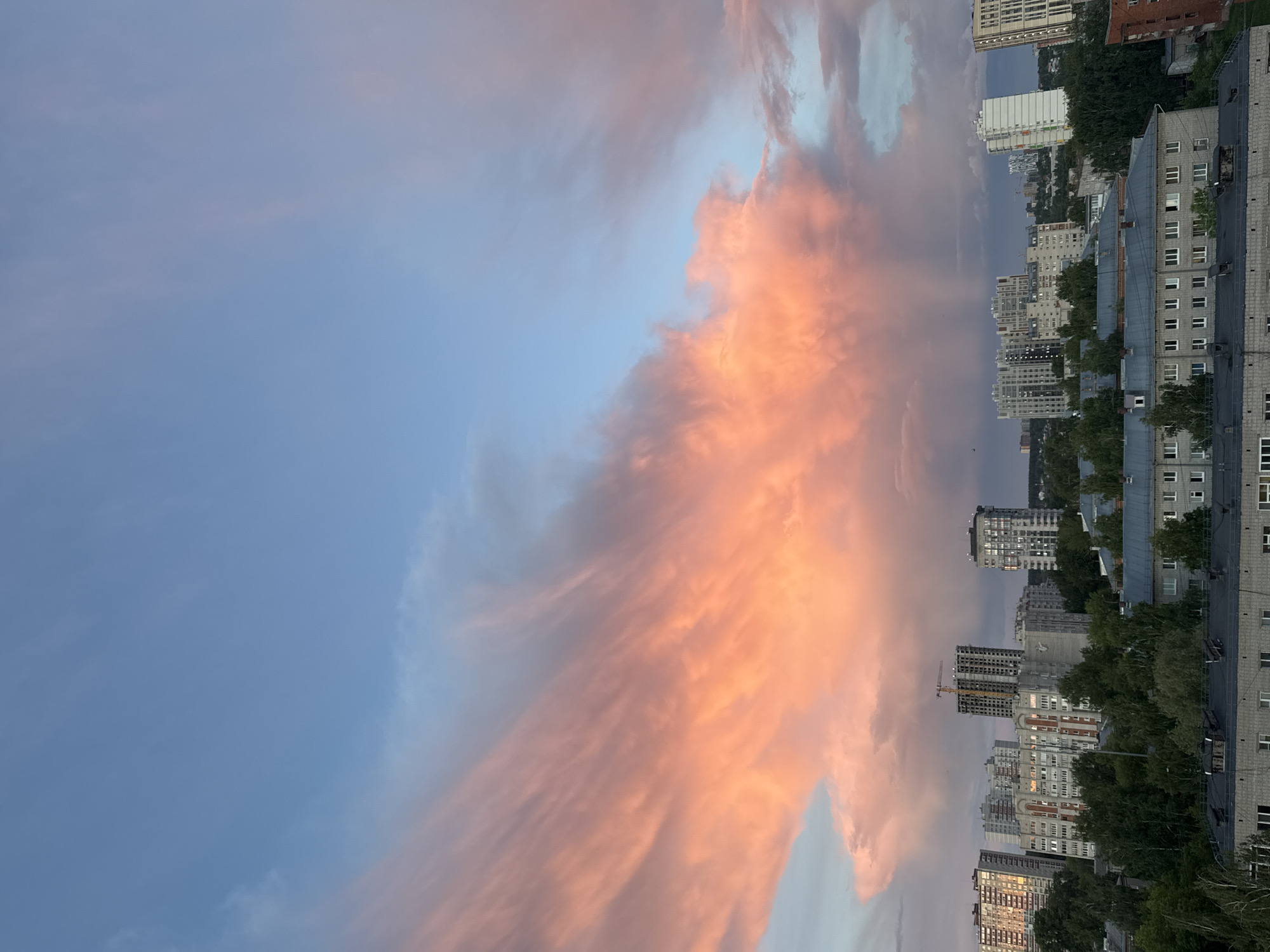The image shows a view in Izhevsk, Russia, at sunset. The sky is filled with long, orange-pink clouds contrasting against the blue sky. In the foreground, there are buildings of varying heights. Trees are interspersed between the buildings. A construction crane is visible among the buildings. The photo is taken at an angle.