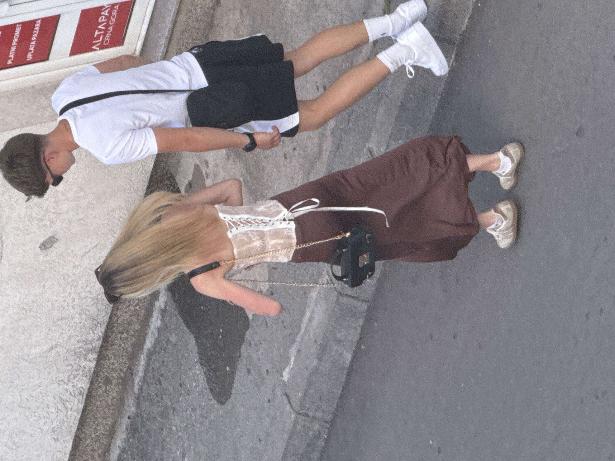 The image shows two people, a man and a woman, lying on their backs on a paved surface. The man is wearing a white t-shirt, black shorts, and white sneakers. He has dark hair and is wearing sunglasses. A black bag is slung across his chest. The woman is wearing a white lace top and a long brown skirt. She has blonde hair and is wearing sneakers and white socks. She is carrying a small black bag with a gold chain strap.
They appear to be relaxing outdoors, possibly on a street or walkway. The background suggests an urban environment with a textured wall on the left and a grey paved ground. There is some text visible on a sign in the background, which appears to be in a foreign language. The lighting suggests it is daytime, with bright sunlight creating shadows. The overall mood seems relaxed and casual.
The visible text on the sign reads "PLATNI PROMET", "UPLATA PAZARA", and "ALTAPAY CRNA GORA". This indicates a commercial area, possibly a payment or currency exchange point. Given the location context of Budva, Montenegro, it's likely a street scene in this coastal city.