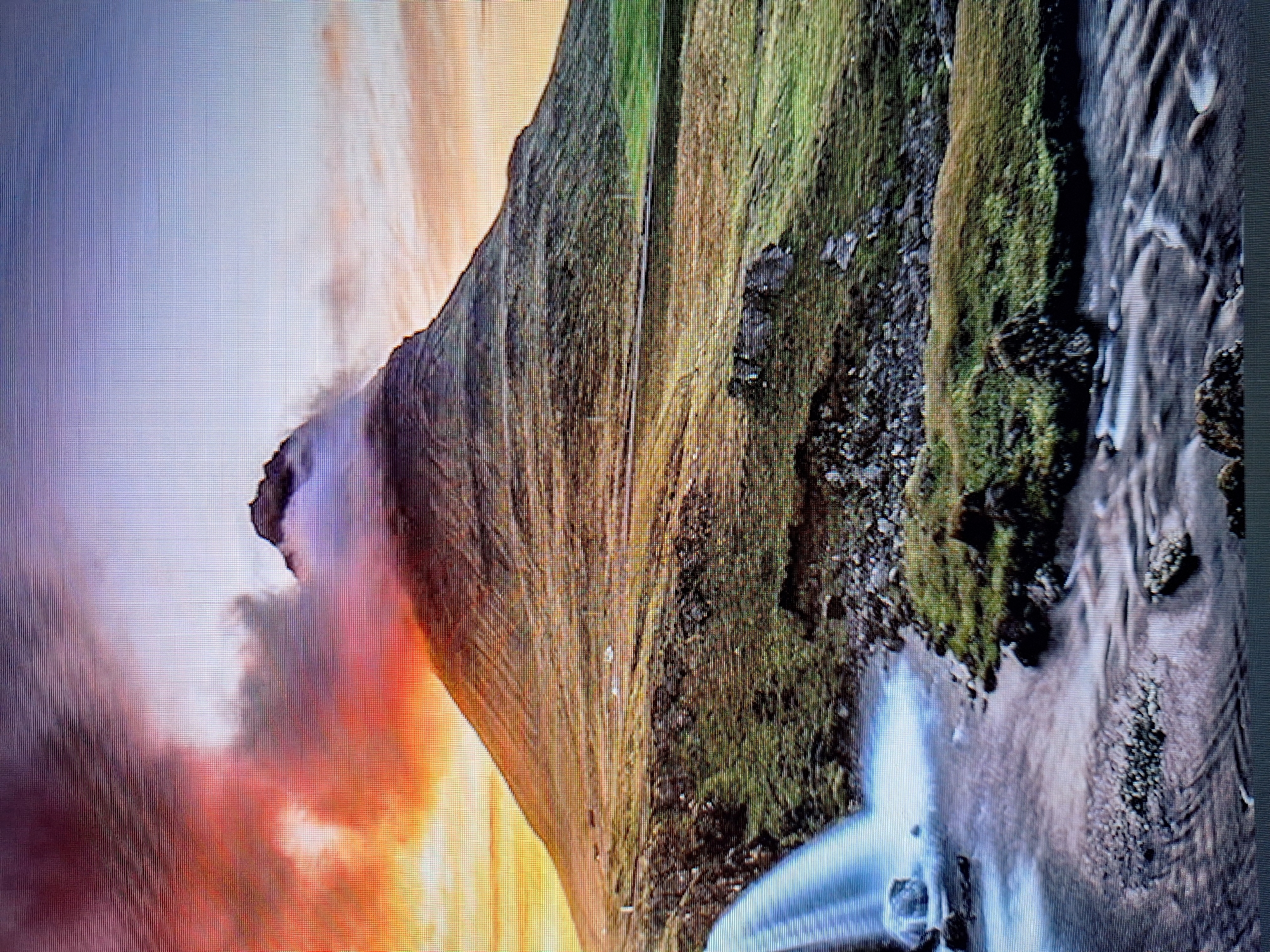 The image displays a mountainous landscape with a waterfall and a winding river. The sky is filled with dramatic clouds, exhibiting hues of orange, pink, and purple, suggesting either sunrise or sunset. The mountain is covered in lush green grass and some patches of rock. The waterfall cascades down a rocky incline into the river, which flows through a rocky bed. The overall scene conveys a sense of natural beauty and tranquility. There is no information within the image that would suggest a location in Varna, Bulgaria.
