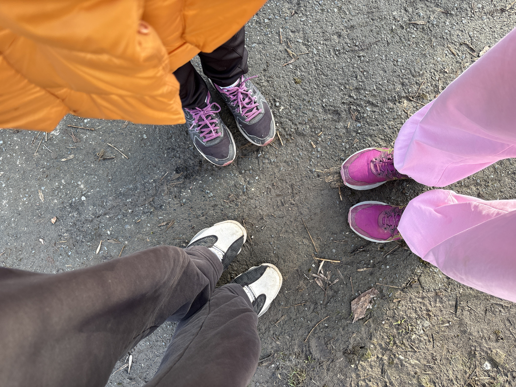 From a high-angle perspective, the image shows the lower portions of three individuals standing in a circle on an outdoor surface of grey asphalt and dirt, likely a path or parking lot.
The person on the upper left is wearing an orange puffer jacket, black pants, and grey running shoes with pink shoelaces. The person on the right is wearing pink pants and magenta running shoes. The person at the bottom of the frame is wearing grey sweatpants and white sneakers with black accents.
The ground has scattered twigs and debris. The scene has natural daylight.