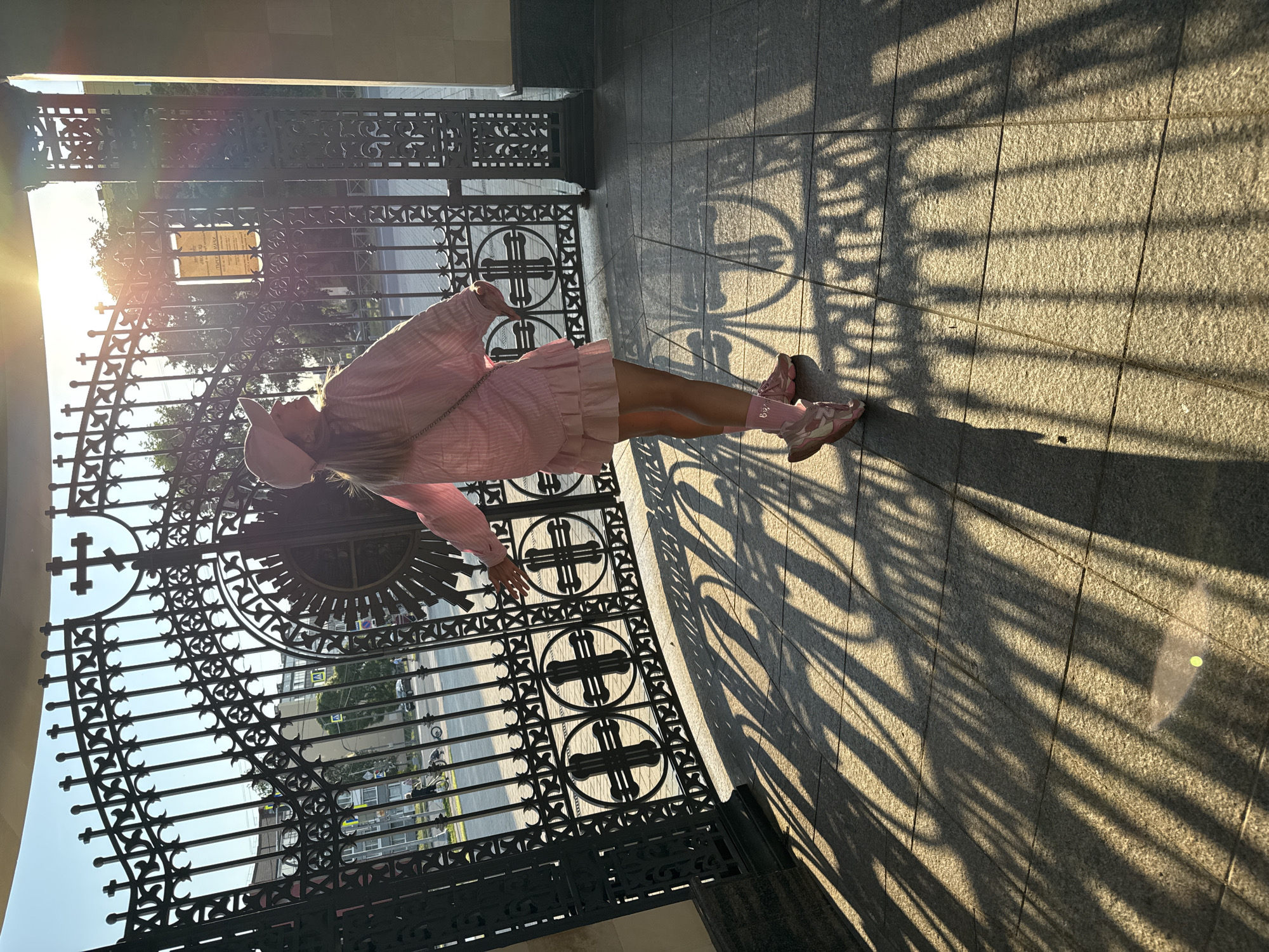 A young woman stands in front of a large, ornate black metal gate, her body turned towards the left side of the frame. She is wearing a pink outfit that includes a long-sleeved top, a pleated skirt, a hat, and pink sneakers with light pink accents. Her socks are also pink. The sun is setting, casting long shadows from the gate onto the granite-tiled ground. The light flares strongly from the left, creating lens flare effects. Through the gate, trees and buildings are visible, as well as a street with cars and a motorcycle. A sign with text is attached to the gate. The overall scene suggests a warm, late afternoon or early evening in a public space, possibly a park or a historical site. The mood appears peaceful and serene, with the golden sunlight creating a soft ambiance. The visible text on the sign is in Cyrillic.