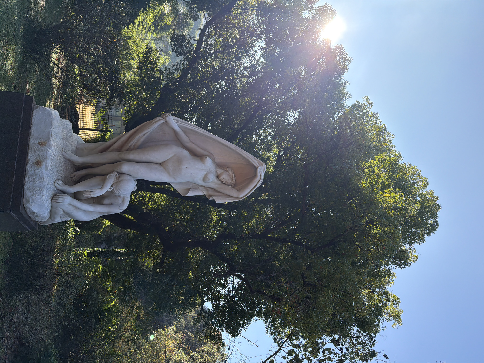 The image shows a marble sculpture in a garden. The sculpture depicts two nude female figures. One figure stands upright, draped in fabric that partially obscures her body, while the other figure kneels before her, head bowed. The sculpture is set against a backdrop of lush greenery and a clear blue sky. The sun shines brightly, creating lens flare and highlighting the leaves of a large tree. This sculpture is located in the Rosedal de Palermo, a public rose garden within the larger Parque Tres de Febrero in Buenos Aires, Argentina.