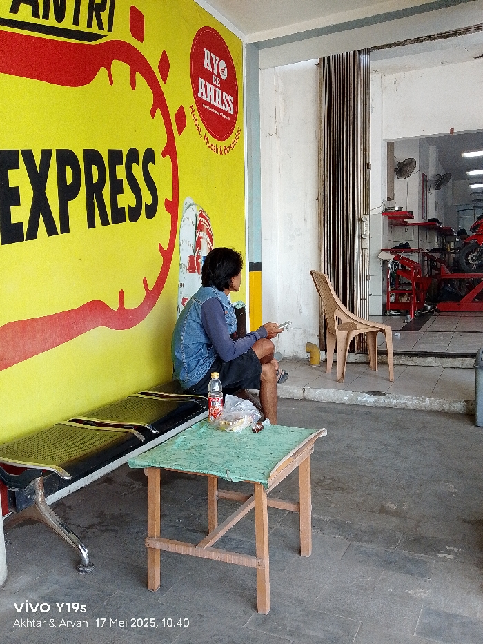 The scene is located in Gegesik, Indonesia at the coordinates -6.536 latitude and 108.432 longitude. A man wearing a blue denim jacket and dark blue pants sits on a black bench in front of a yellow wall with the words "EXPRESS" in black. The wall is plastered with a red and yellow image that reads "Ayo ke Ahass, Hebat, Mudah & Bergaransi". The man is looking at a phone in his right hand while he holds a bottle of water in his left hand. A small table with a green top and four wooden legs sits in front of the man. A chair is positioned to the right, partially visible. There is an open doorway on the right side of the frame with a small space inside. The interior of the space is cluttered with various objects, including what appears to be a motorcycle tire. The space is bright with light coming in from the front of the building. The floor is grey tile. The space has a small table and a chair. The scene is shot with a slightly tilted perspective. There is no evidence of any illegal activity occurring in the scene.