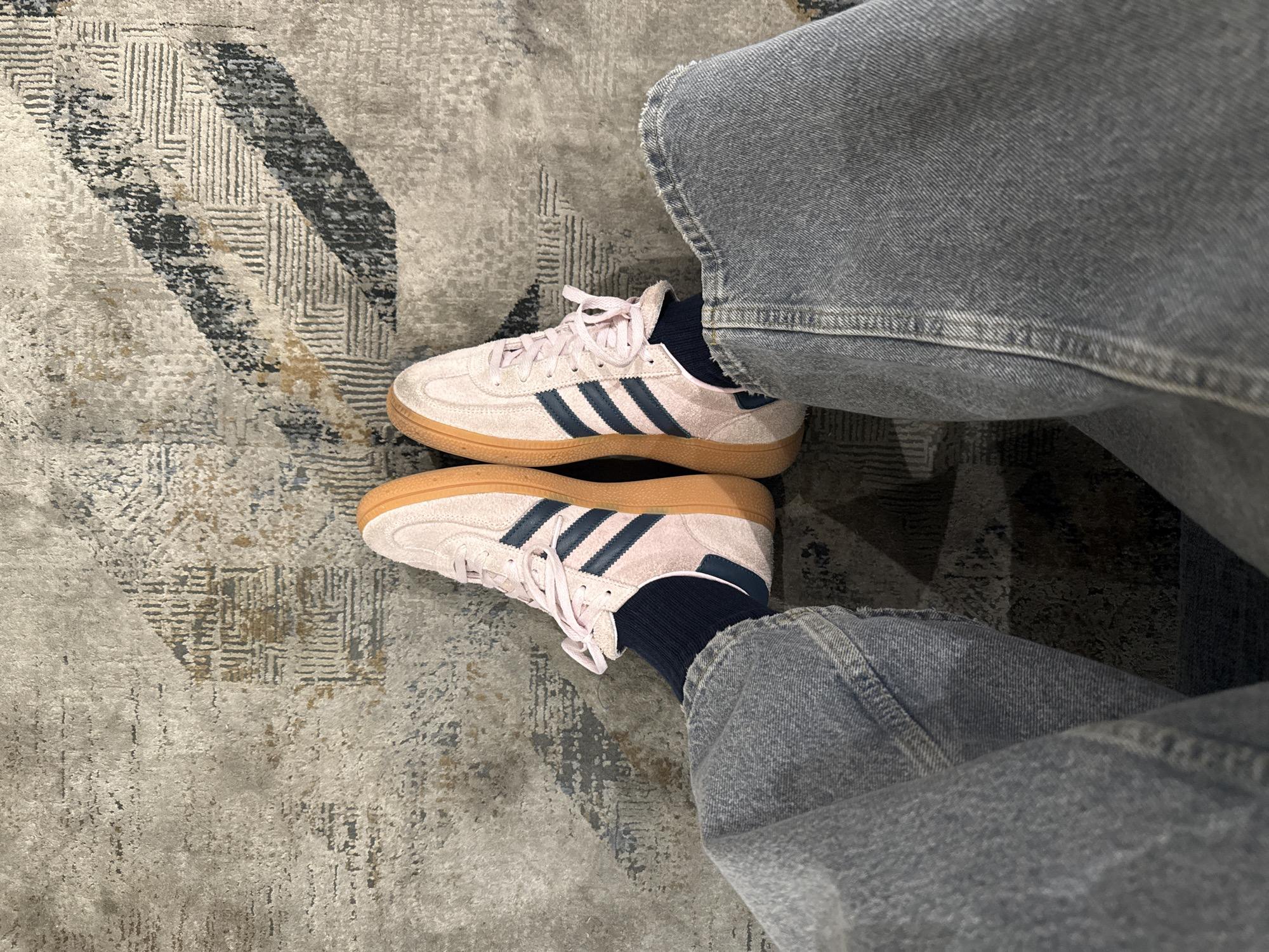 The image shows a person wearing light gray jeans and light pink Adidas sneakers with navy blue stripes. The person is wearing dark blue socks. The shoes are on a gray patterned rug. The location is unclear, but the picture was taken in Mexico City, Mexico.
