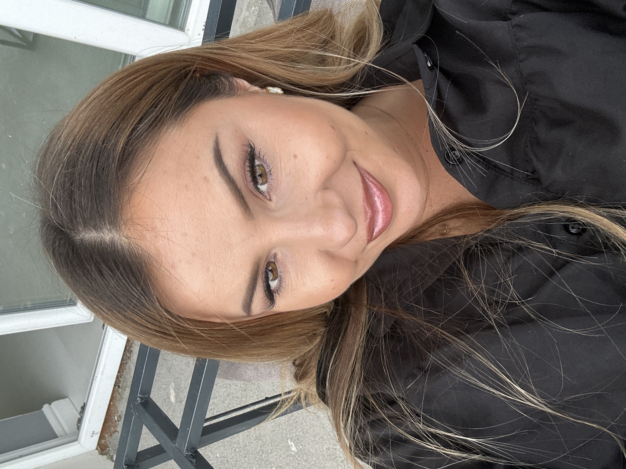 A close-up selfie shows a woman with medium-length blonde hair and brown eyes, wearing a black collared shirt. She has on makeup, including mascara, eyeliner, eyeshadow, and lip gloss. She is also wearing hoop earrings and a delicate necklace. The background appears to be an outdoor setting with a black metal railing and a window frame visible. The location is the city of Asenovgrad, Bulgaria, as indicated by the metadata.