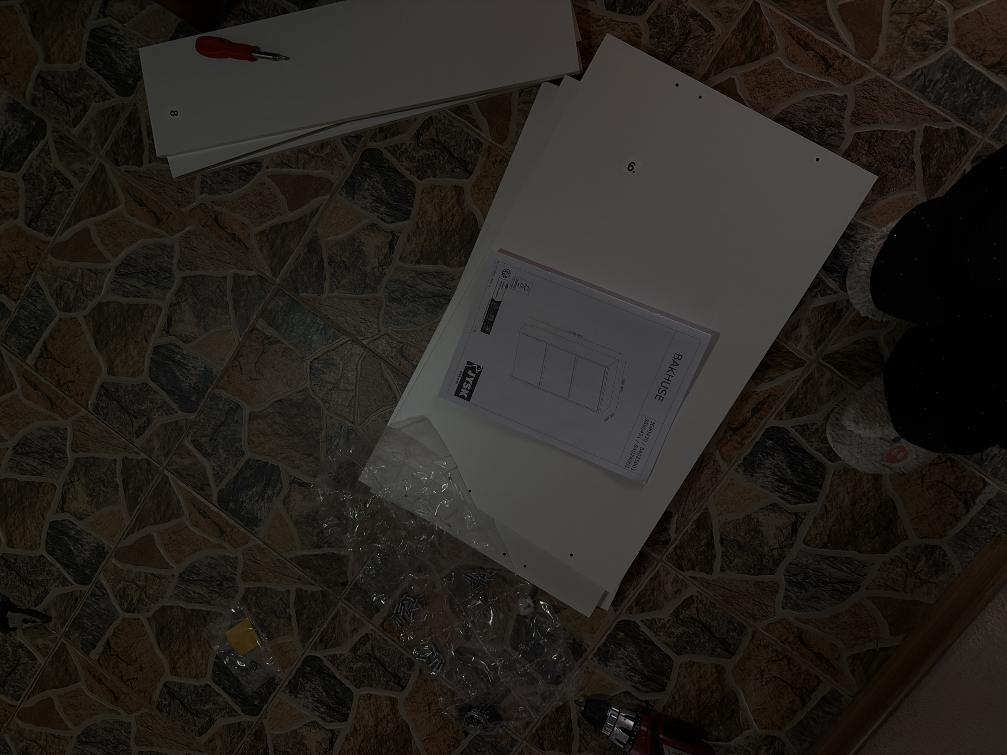A high-angle shot depicts components for assembling furniture on a tile floor. Visible are white furniture panels, labeled "8" and "6," along with an instruction manual from JYSK with the name "BAKHUSE" printed on it and the measurements 1239 mm and 650 mm. A plastic bag of screws and small hardware, a yellow packet, and tools like a screwdriver with a red handle and a drill with a red grip are scattered around. In the top right, a portion of a person's legs dressed in black leggings and white socks are visible. The patterned tile floor features a mix of brown, beige, and gray tones. The setting seems to be indoors, potentially during the assembly of new furniture. The location in the city of Sofia, Bulgaria is indeterminate from the image.
